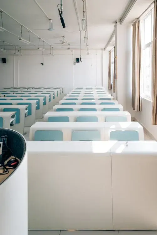 Bright white classroom with rows of desks featuring turquoise chairs, pendant lighting, and large windows