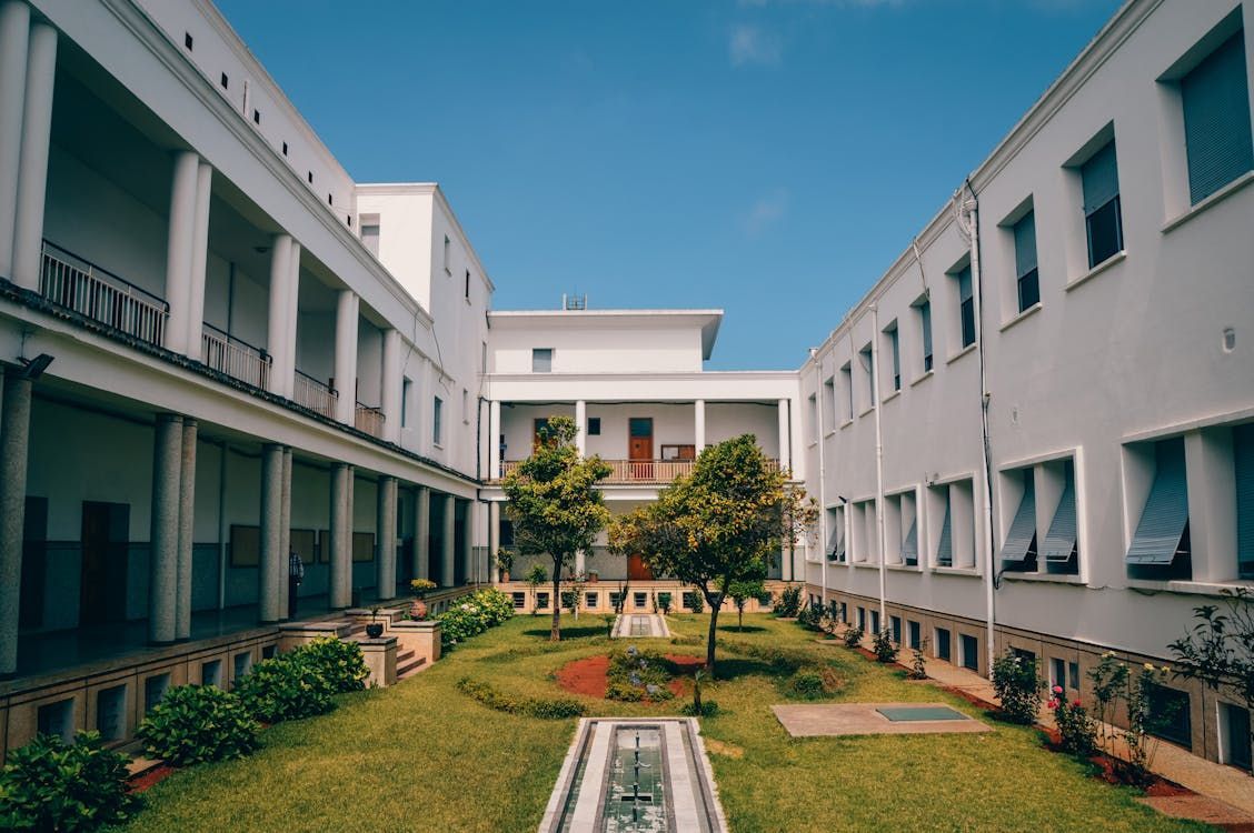 School courtyard with white buildings on three sides, central lawn area with trees, and geometric garden design