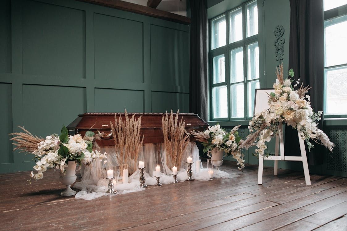 Memorial service setup with wooden casket surrounded by white flowers, silver candles, and silk draping in green-walled room