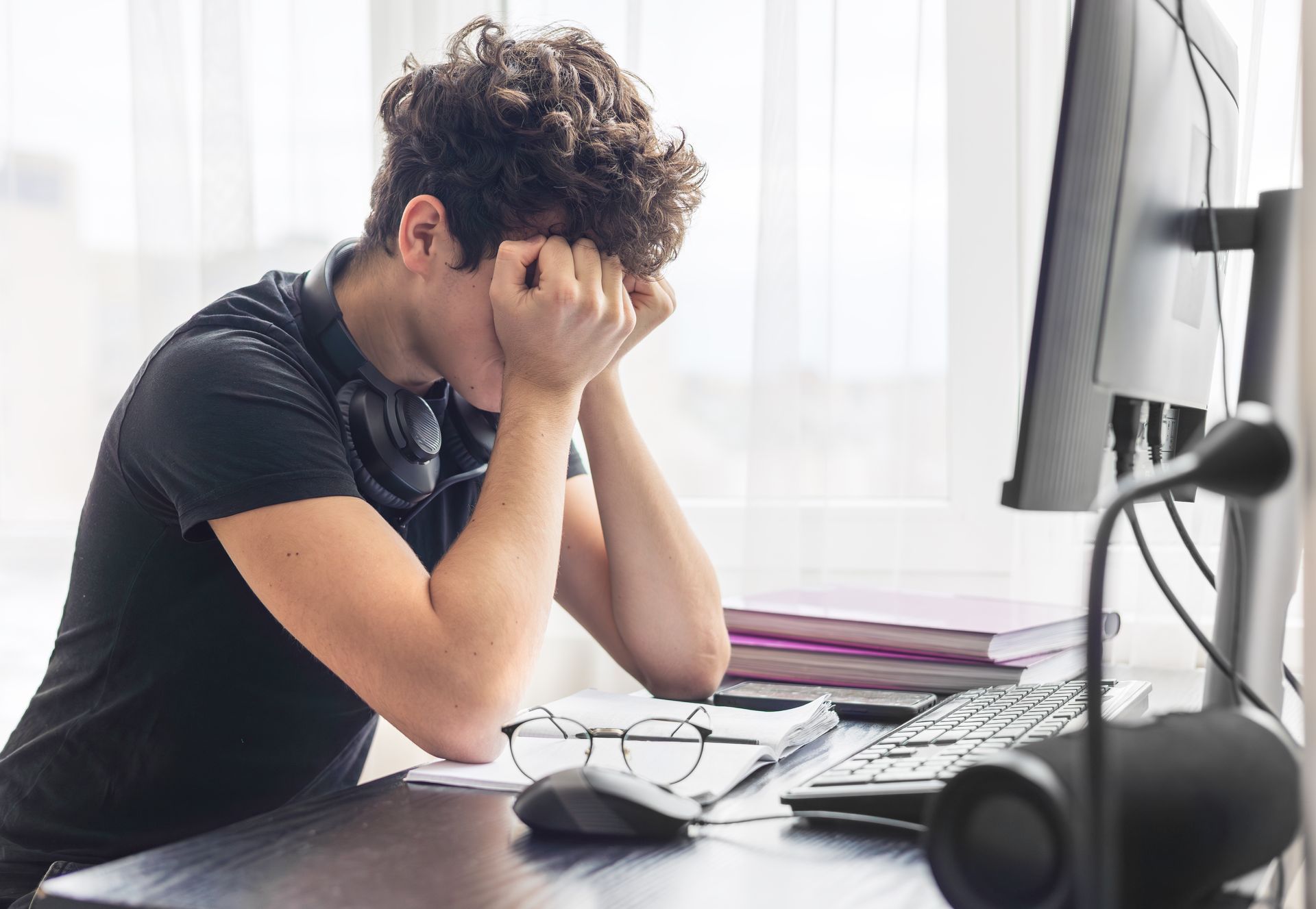 A person sits at a desk with their head in their hands, looking frustrated or exhausted while working on a computer.