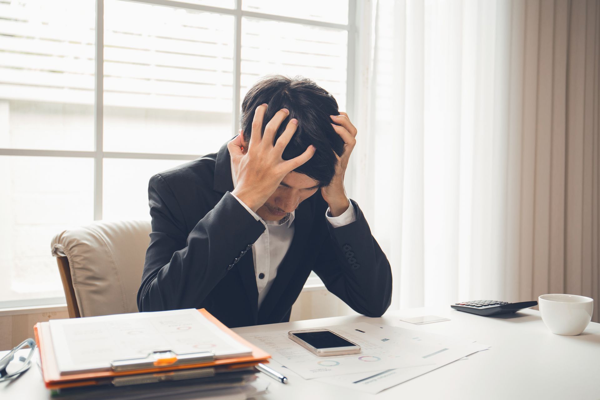 A person in a suit sits at a desk, head in hands, looking stressed while surrounded by documents and a smartphone.