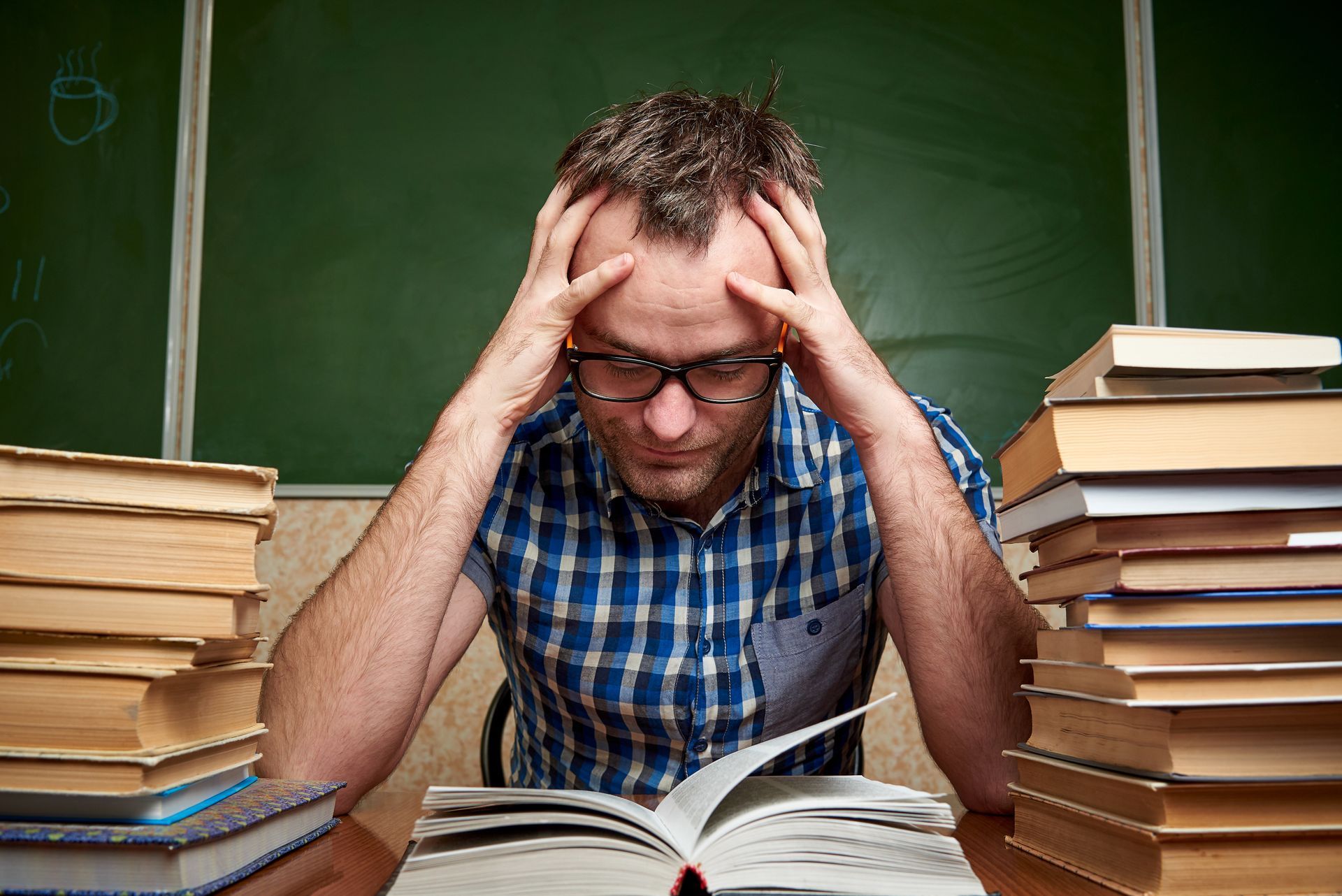 A person with glasses in a blue plaid shirt sits at a desk, stressed, holding their head with two large stacks of books.