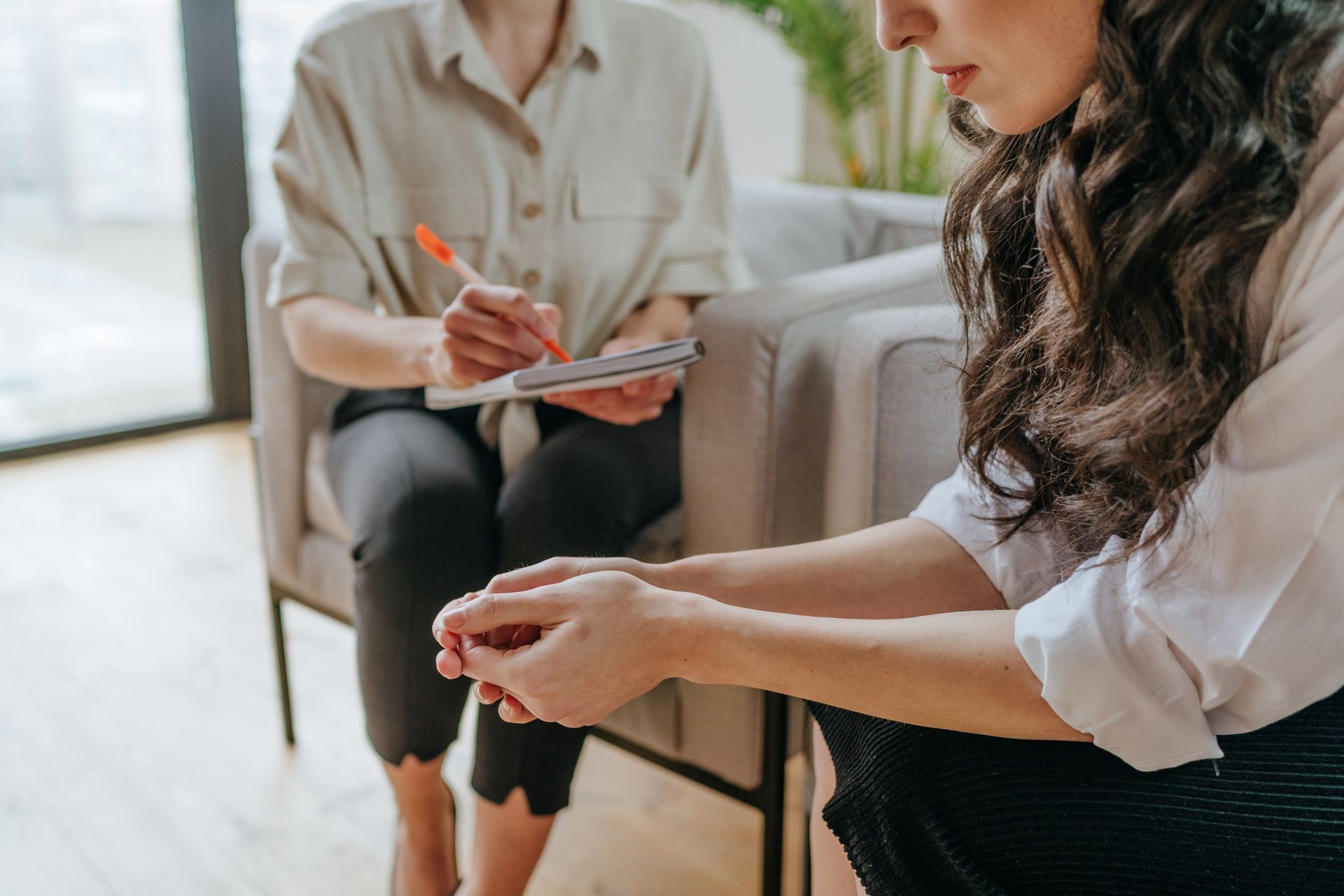 A person sits in a chair taking notes while another person sits opposite, hands clasped, during a professional meeting.