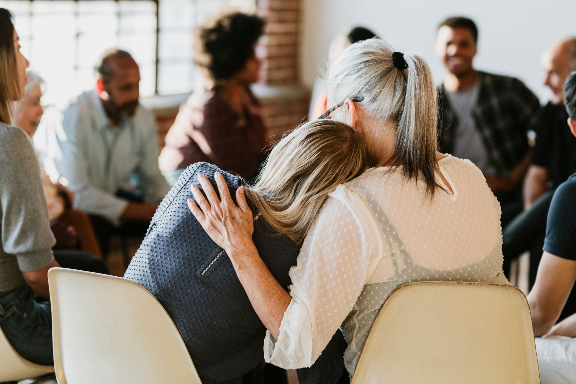 A support group session where one person leans their head on another's shoulder, who is holding them in a supportive embrace.