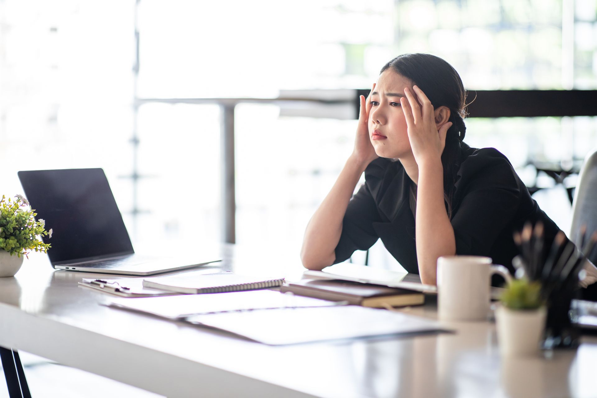 A person in a black shirt sits at a desk with a laptop and documents, holding their head with a stressed expression.