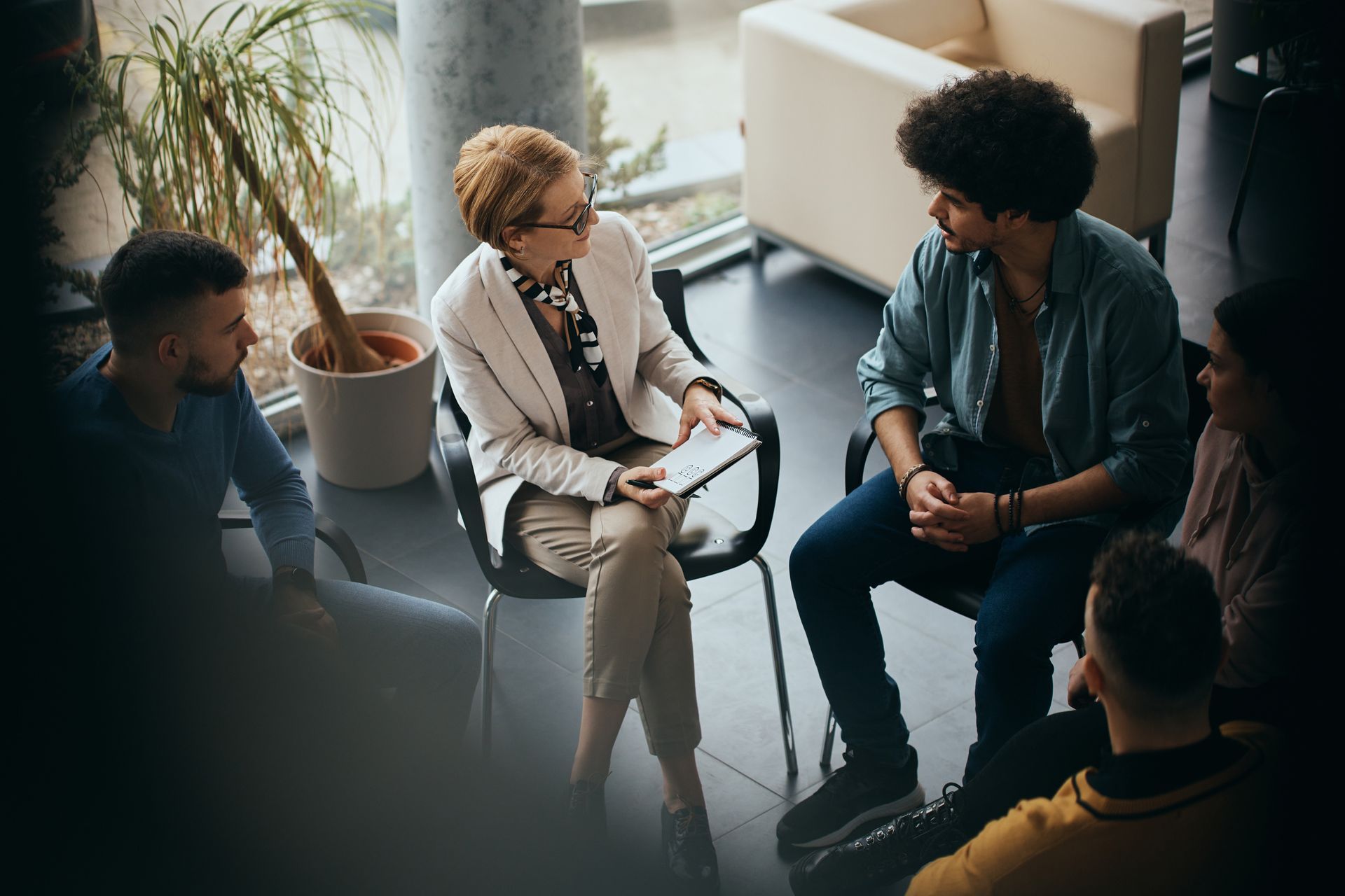 A professional in a white blazer talks to a group seated in a circle in a sunlit office.