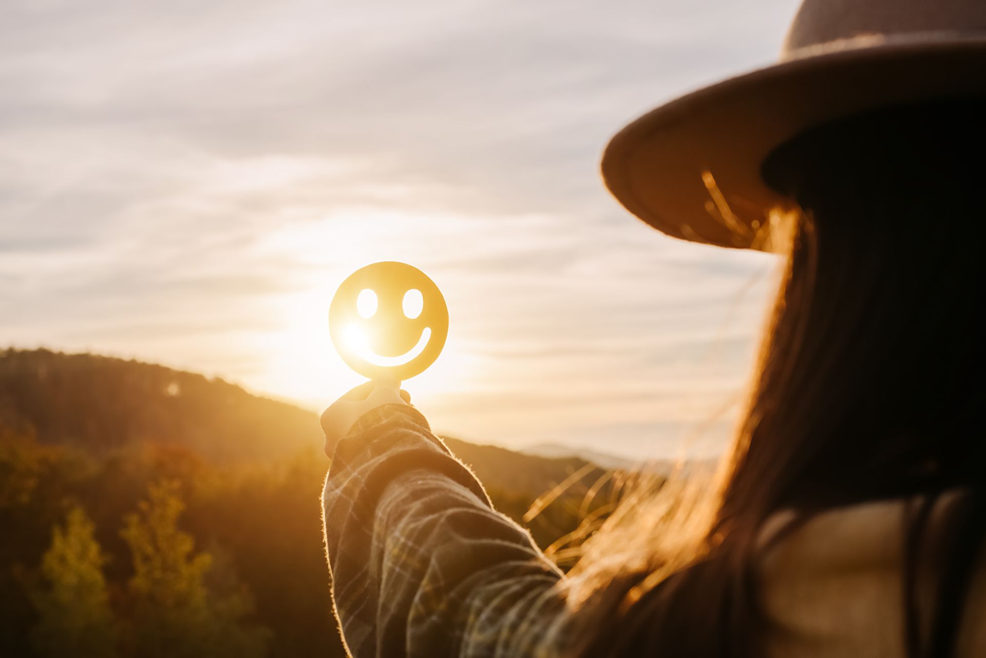 A person holds a small, cutout smiley face against a golden sunset over a hilly landscape.