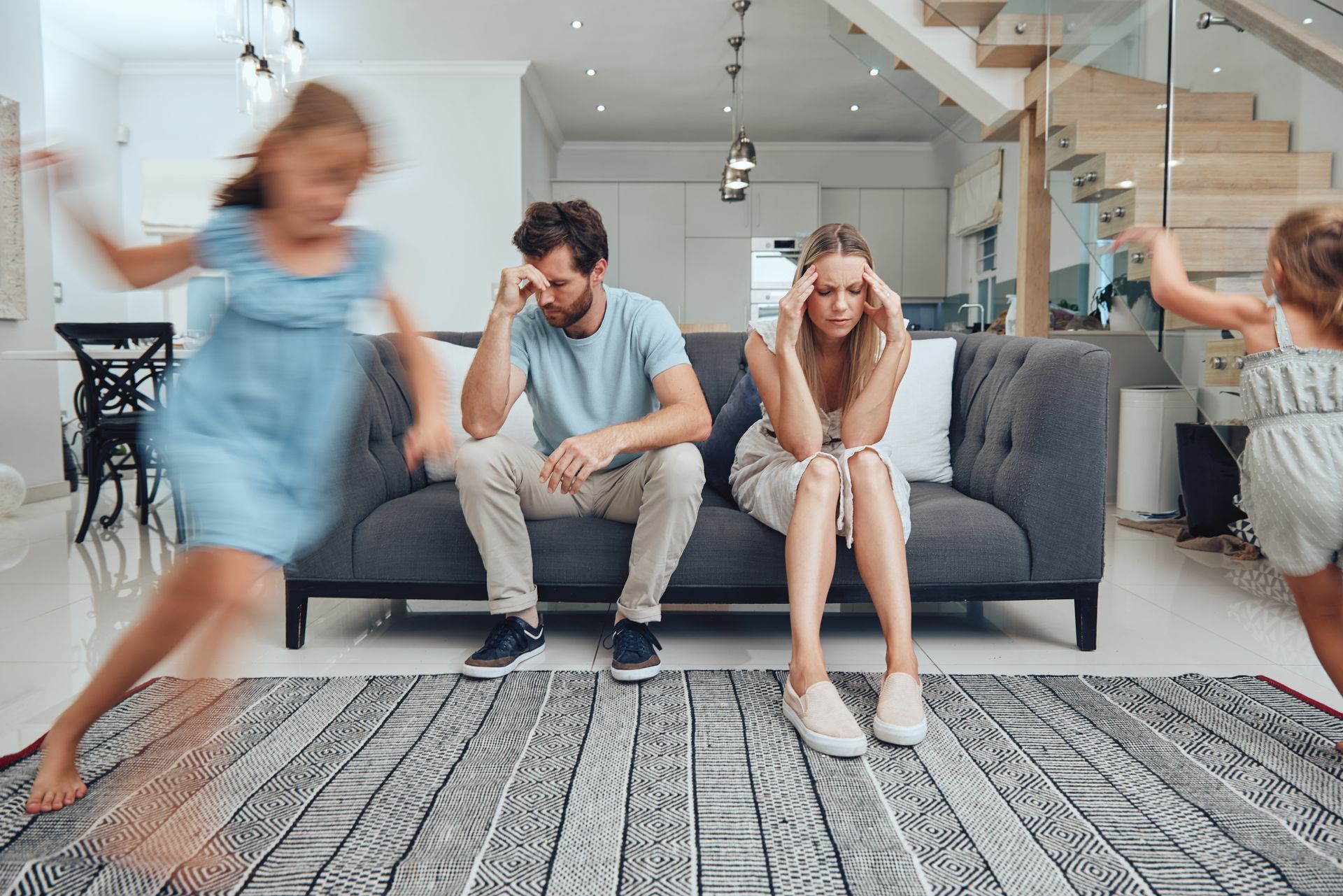 Stressed parents sit on a sofa in a living room while two children play and run around them in a blur of motion.