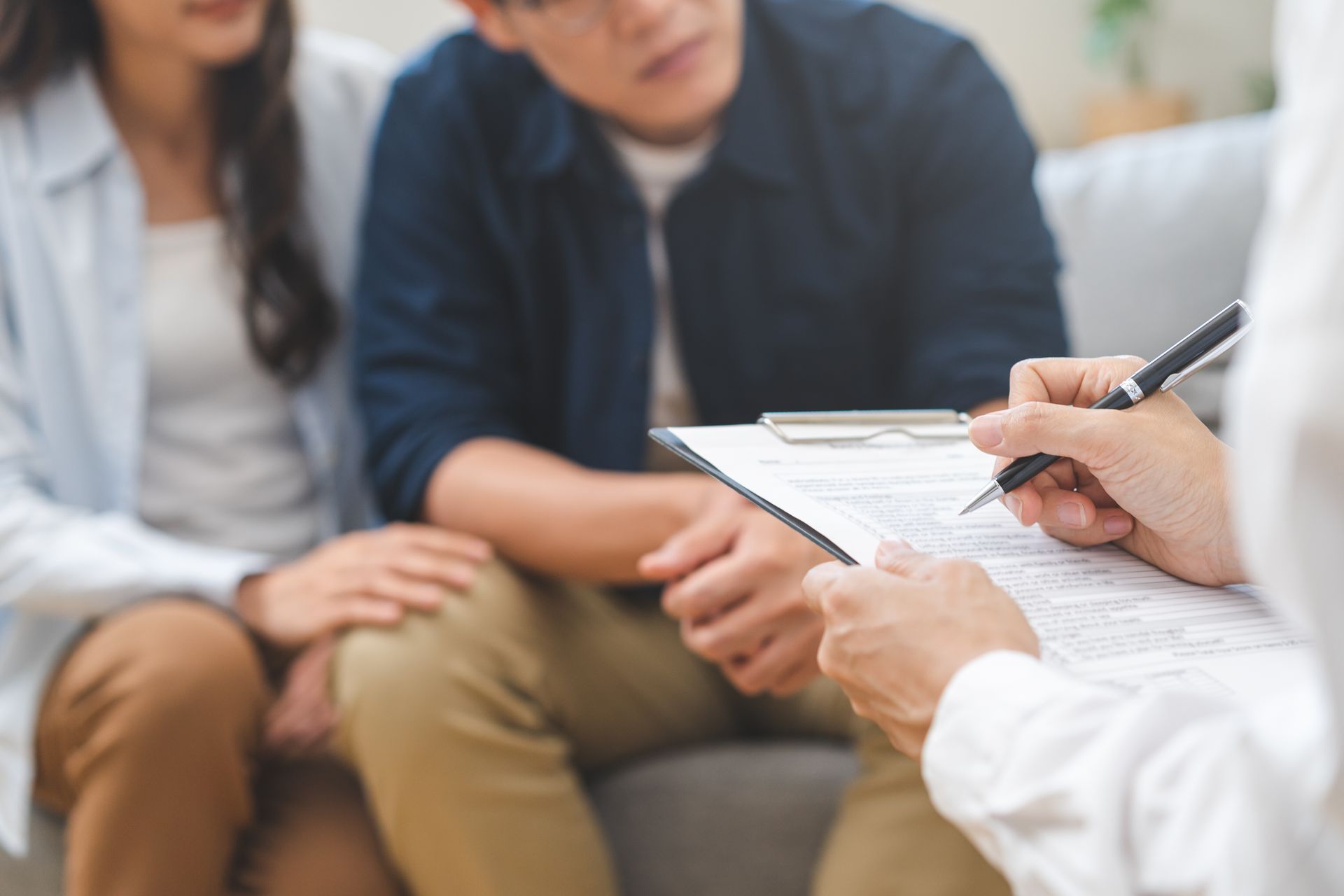 A professional takes notes on a clipboard while sitting with two people during a consultation in a brightly lit room.