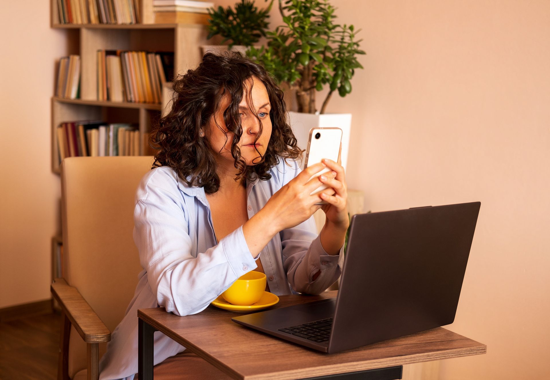 A person with curly hair sits at a desk with a laptop and a yellow mug, looking intently at a smartphone.
