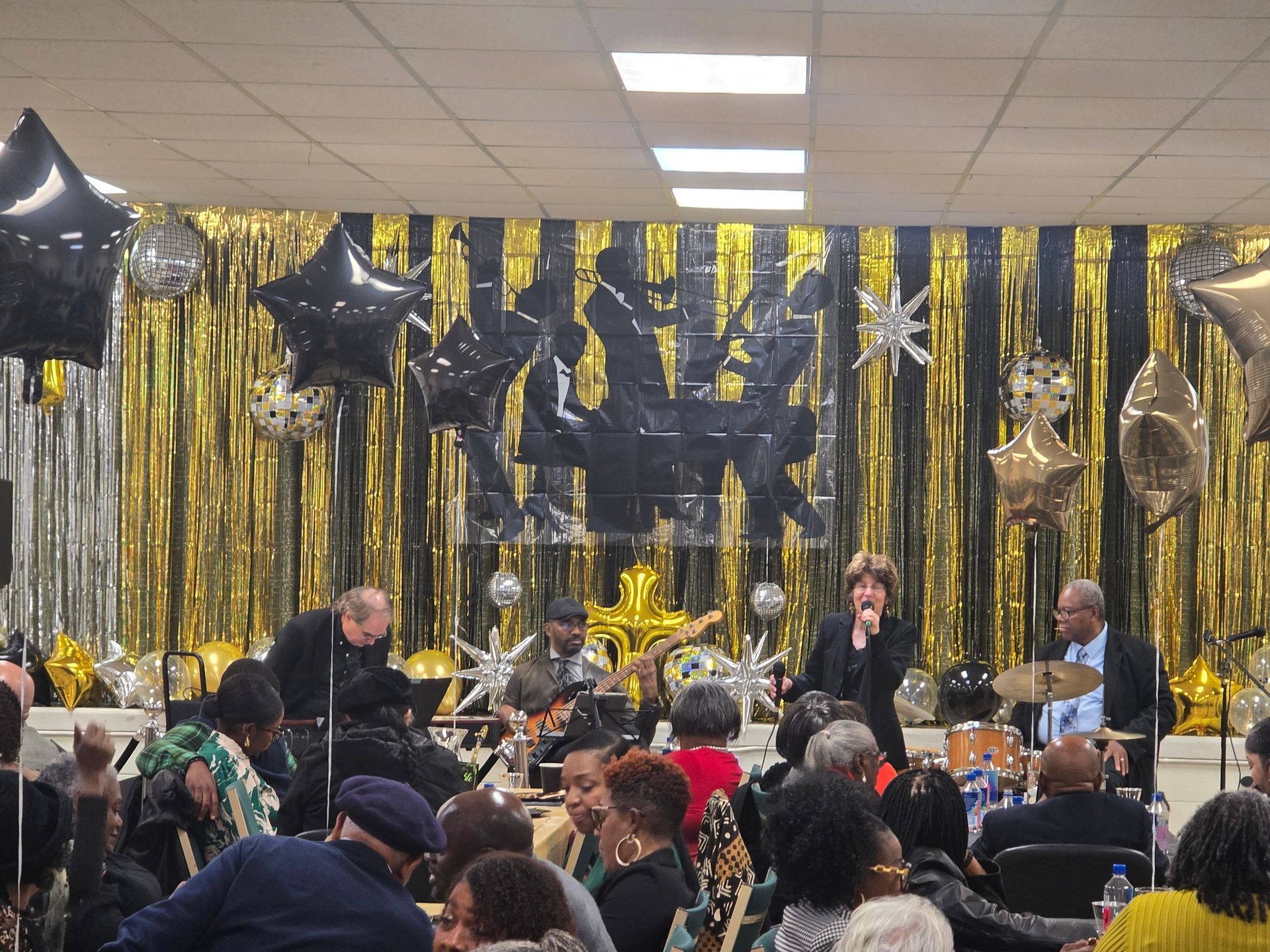 Band performing on stage in front of a black and gold curtain. Audience seated at tables, watching.