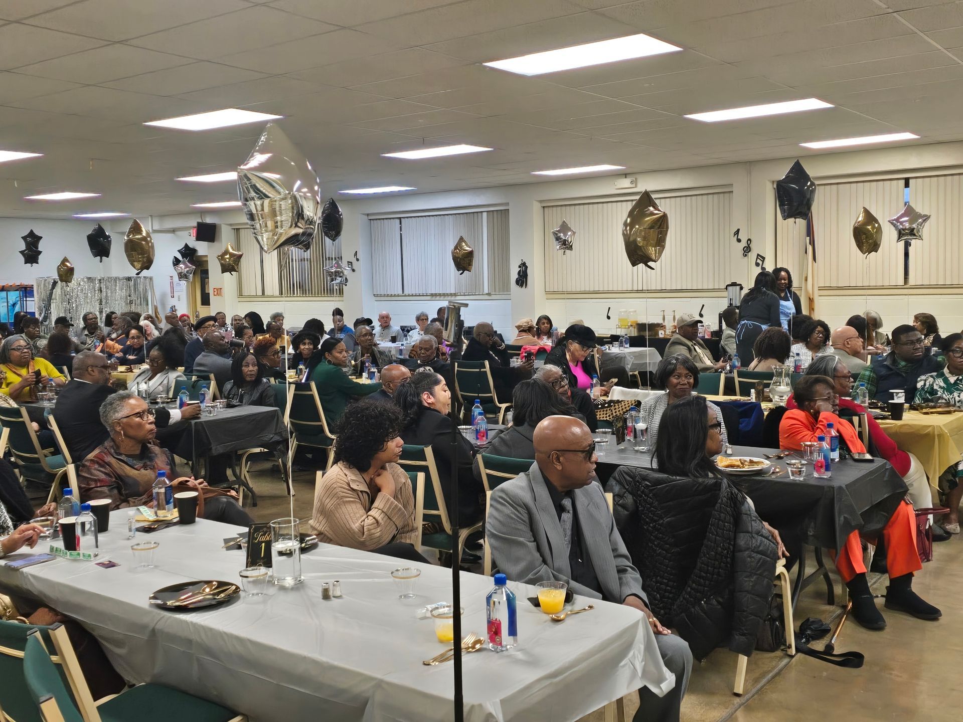 People seated at tables in a decorated hall; attending an event with balloons.