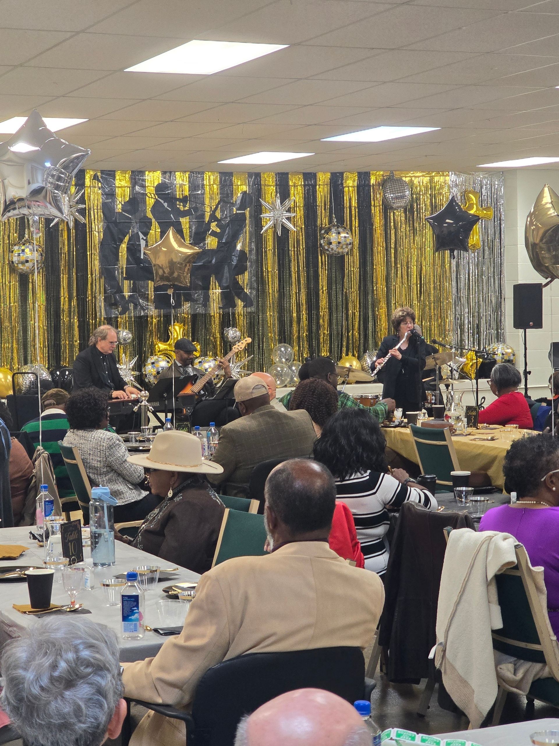 Band playing at a party; gold, black, and silver decorations in background; people at tables watching.