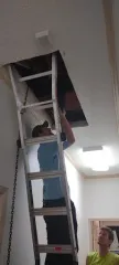 Man on ladder reaching into attic, another man looking up. White ceiling.