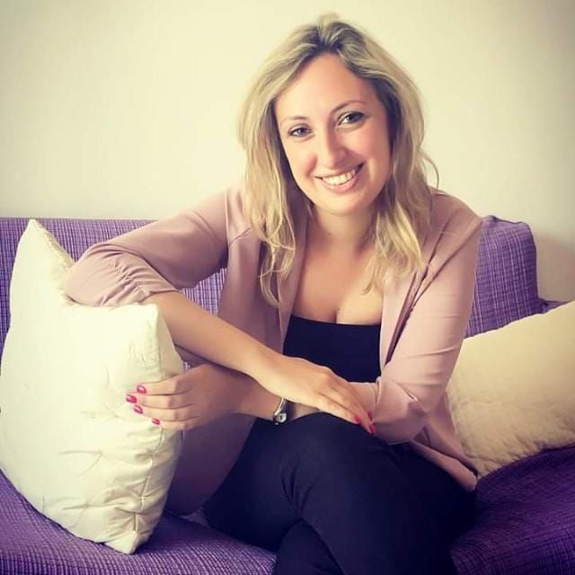 Una donna bionda con una giacca rosa sorride sul divano viola con i cuscini.