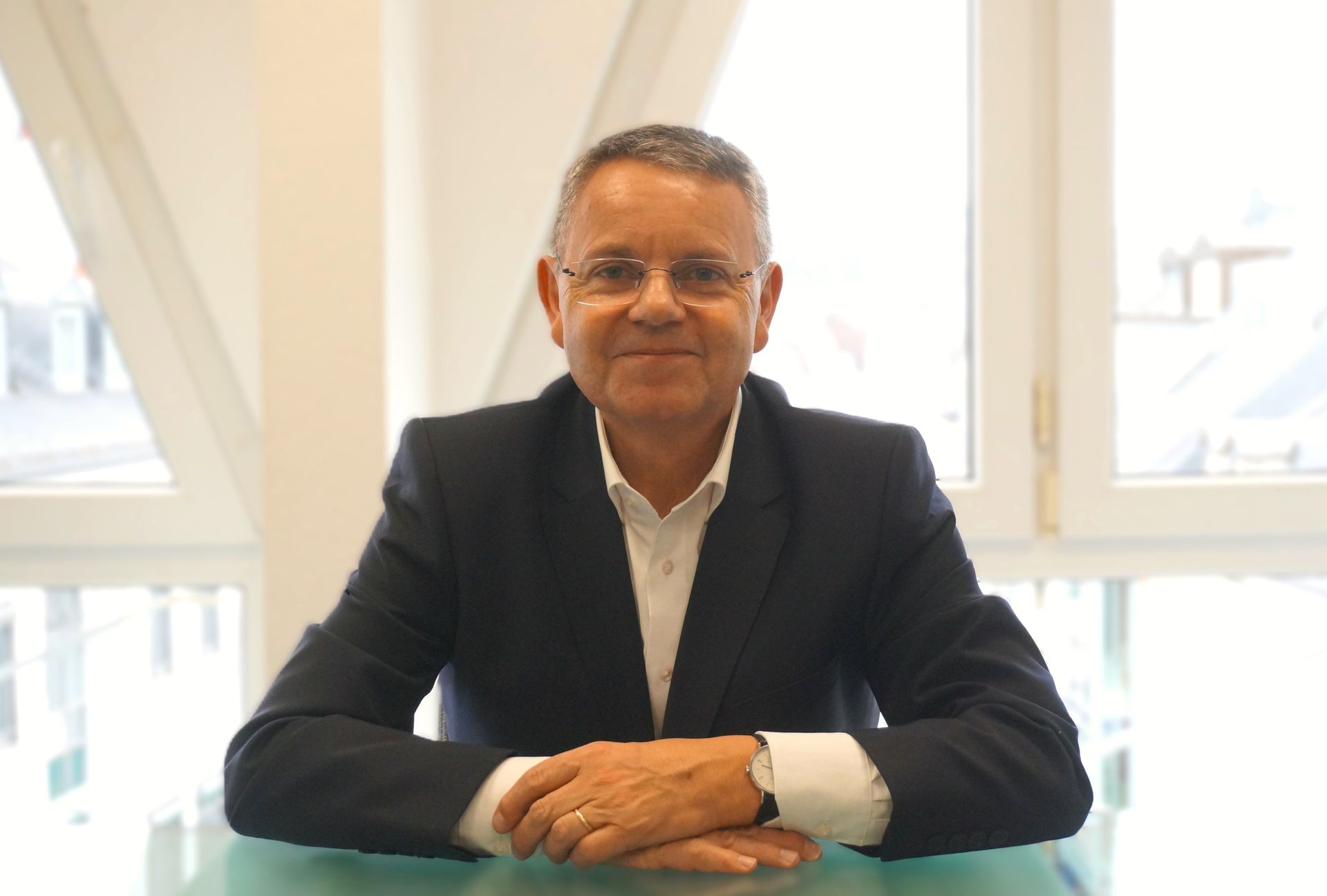 Ein Mann im Anzug sitzt lächelnd an einem Tisch vor einem Fenster.