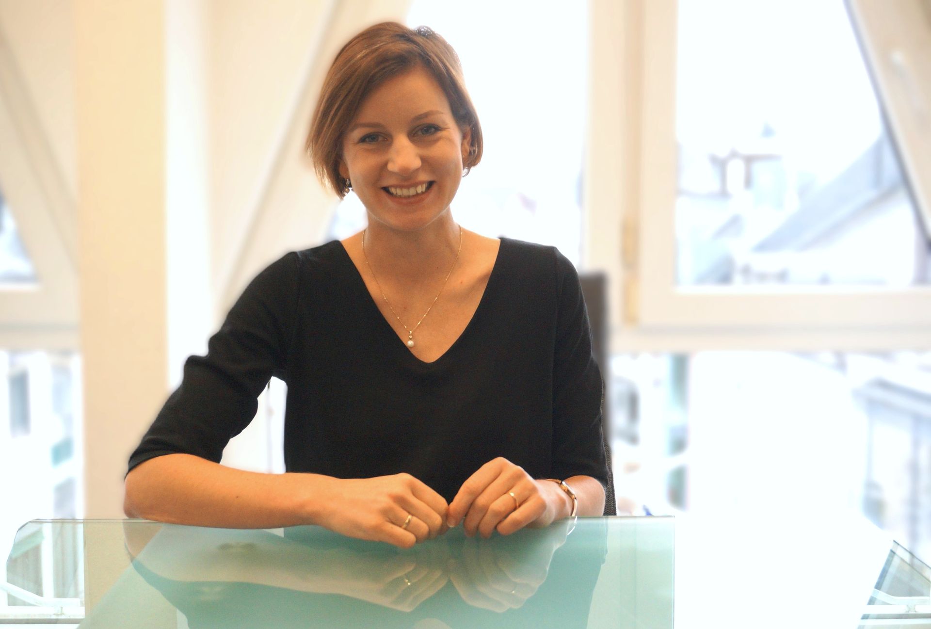 Eine Frau in schwarzem Oberteil lächelt in die Kamera; sie sitzt an einem Glastisch in der Nähe eines Fensters.