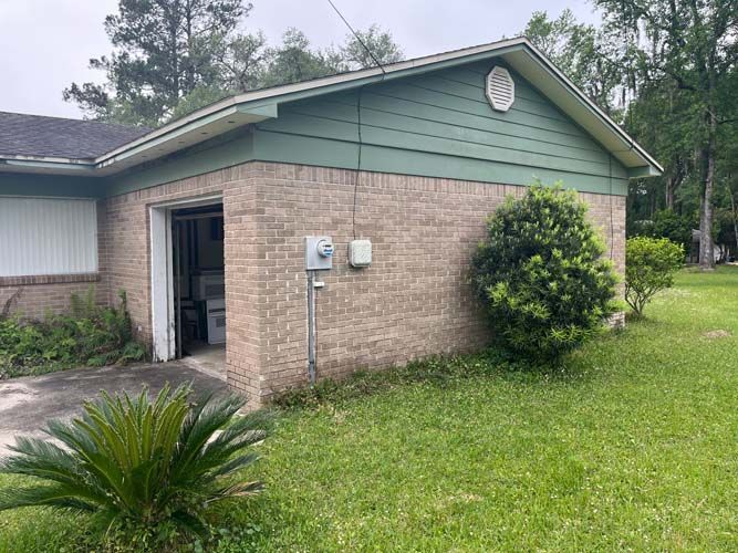 Small beige building with green trim, open doorway, and shrubs beside a grassy lawn.