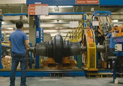 Un hombre con camisa azul observa un gran componente industrial negro suspendido en una fábrica. Se ven maquinaria y equipos.