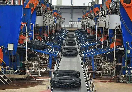 Neumáticos en movimiento por una cinta transportadora en una gran fábrica. Maquinaria azul y naranja rodea la cinta.