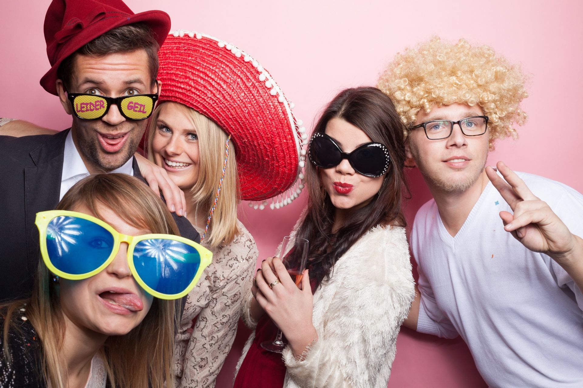 Four friends in a photo booth wearing fun accessories like a sombrero, large sunglasses, and a blonde wig.