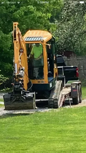 Yellow excavator on grass, operator in cab, parked behind a black pickup truck.