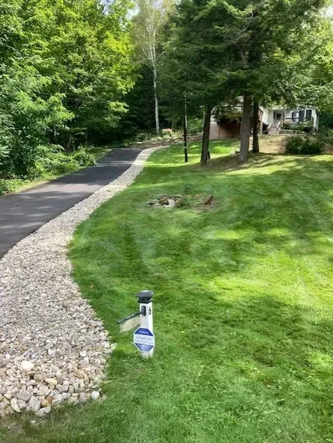 Asphalt driveway bordered by rocks and green grass leading to a house in a wooded area.
