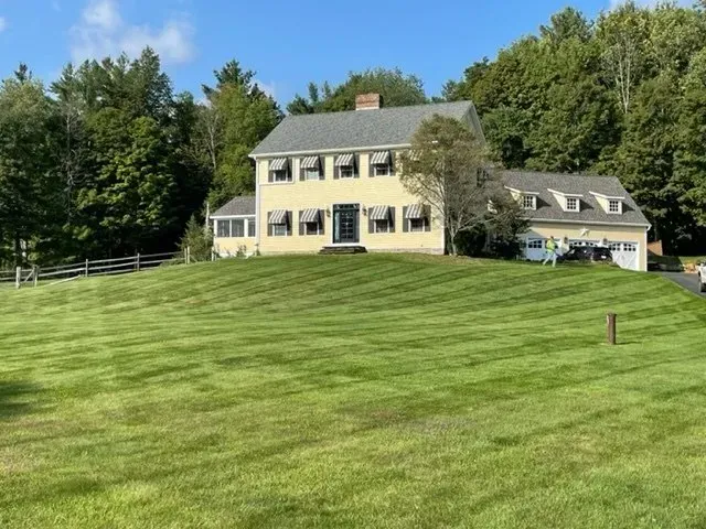 Yellow house on a grassy hill with striped lawn, surrounded by trees under a blue sky.