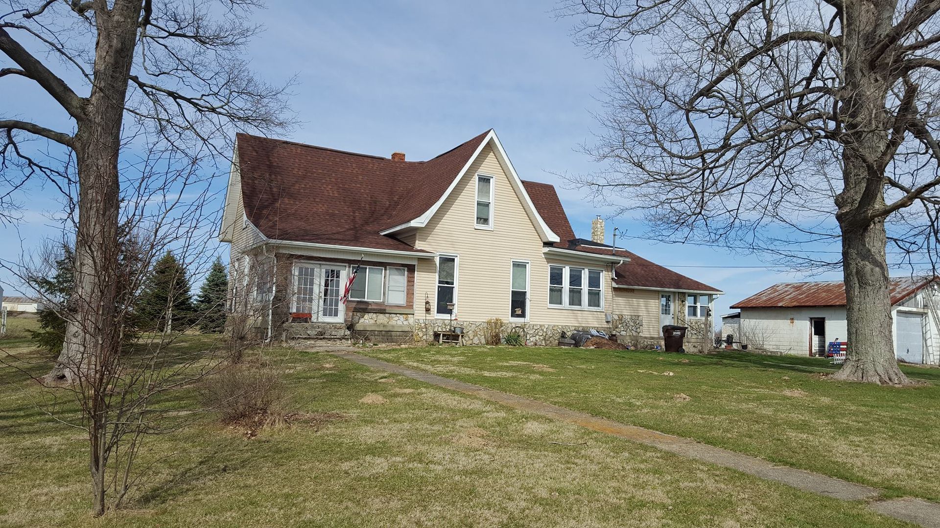 Beige two-story house with brown roof, sitting on a grassy hill, flanked by bare trees and a small outbuilding.