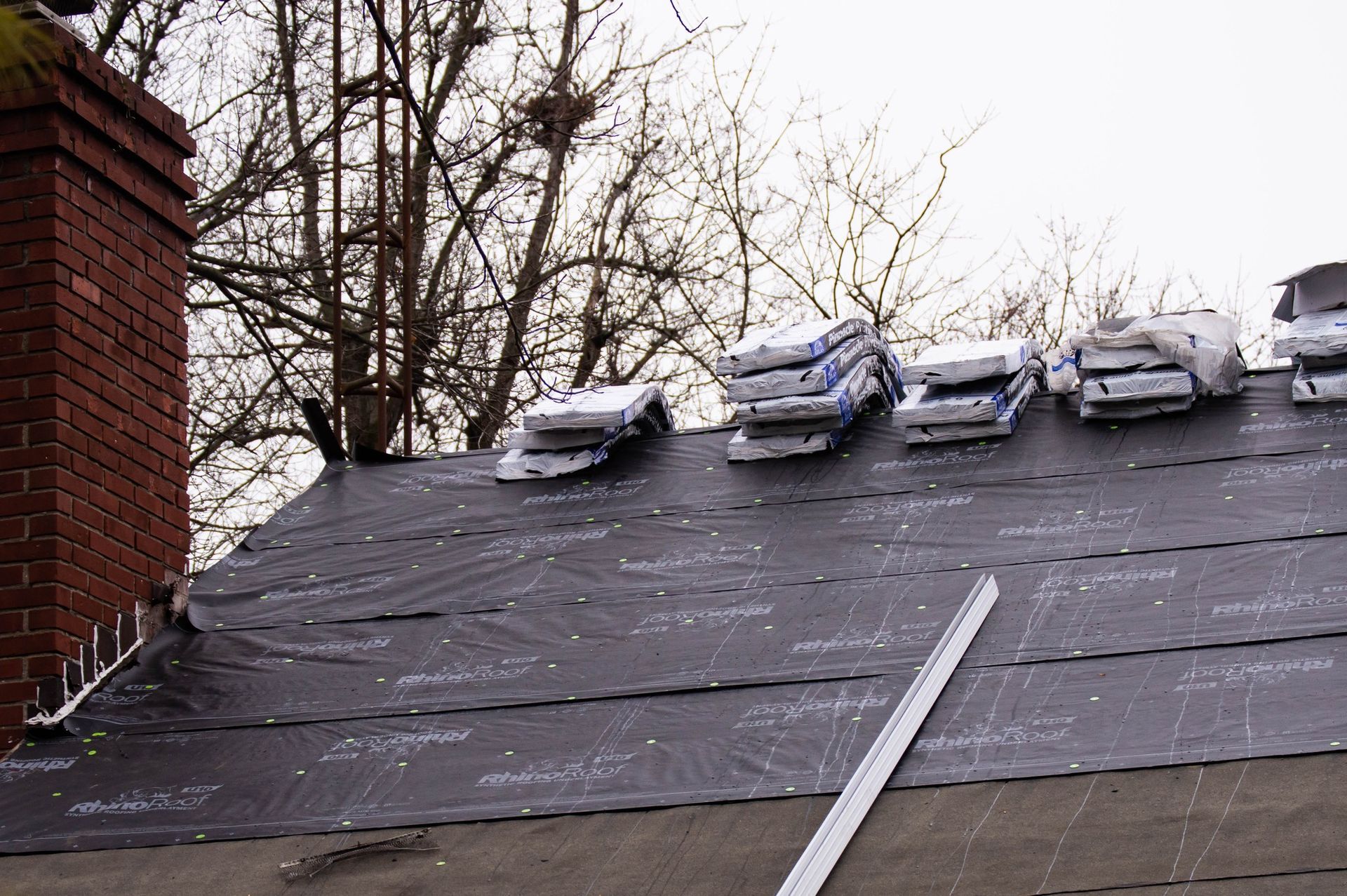 Rooftop with bags of roofing materials next to a brick chimney and bare trees in the background.