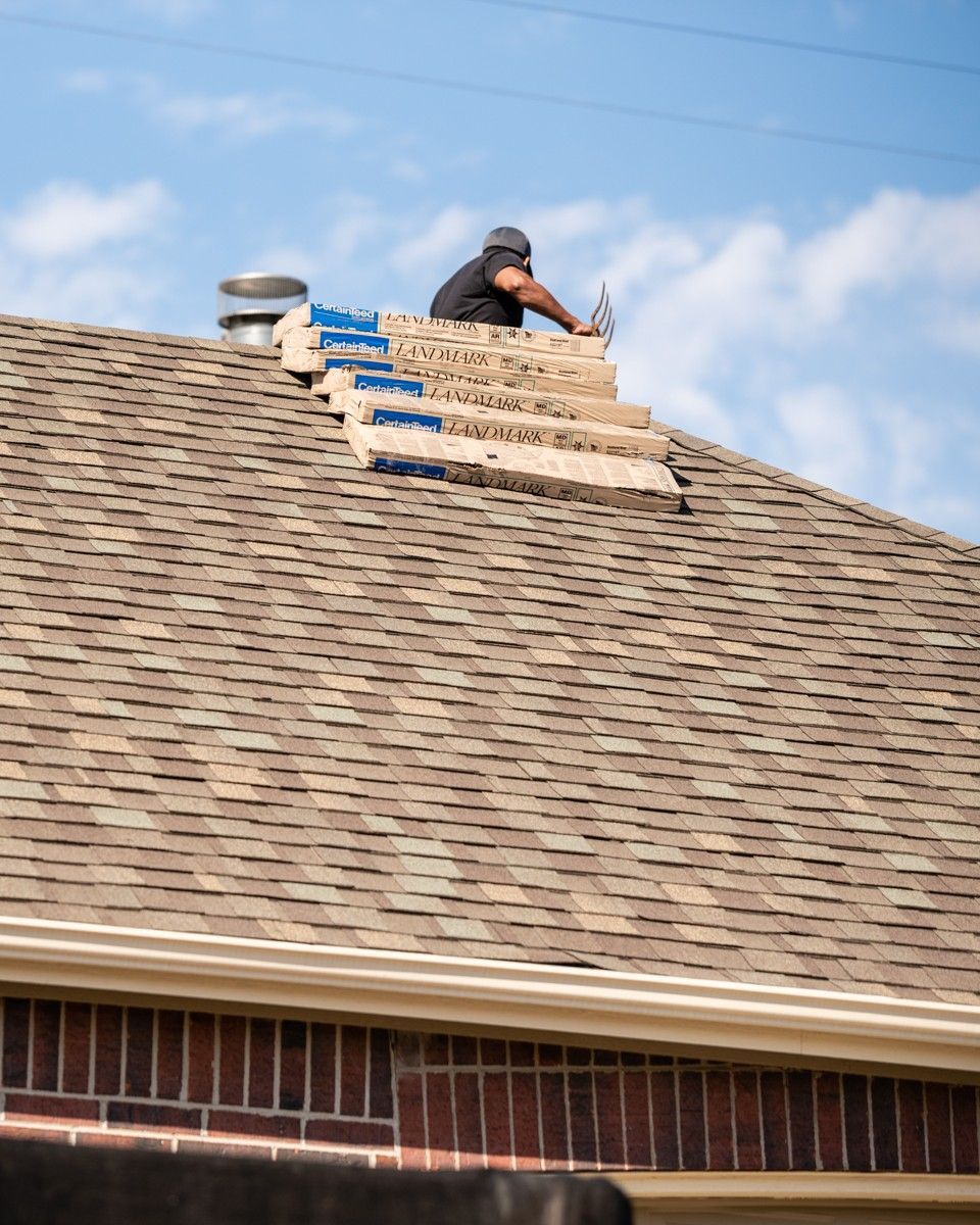 Roofer on a brown shingle roof, working with wooden planks. Sunny day, blue sky, brick building.