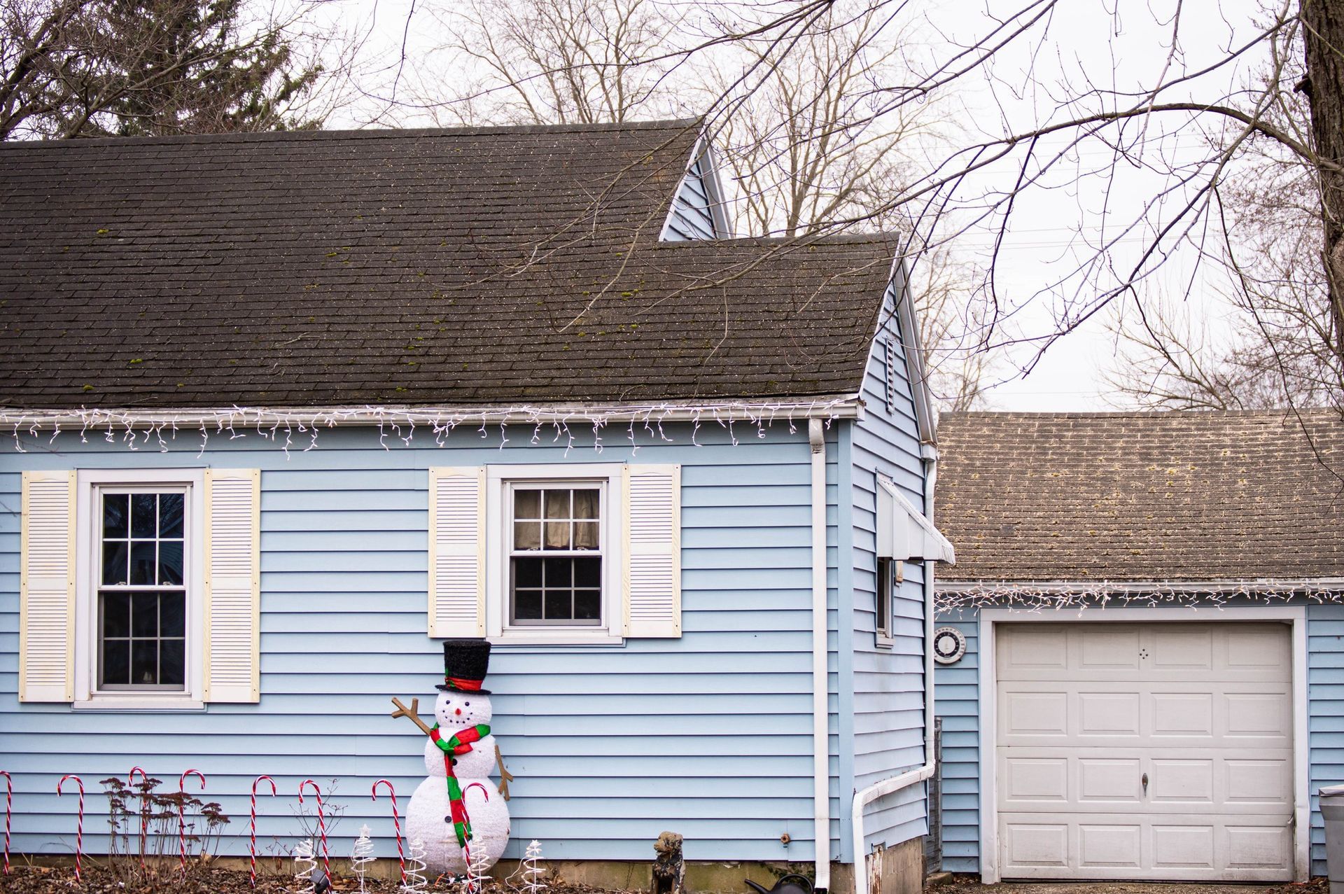 Light blue house with white shutters, a snowman, and a garage in front of a bare tree; Christmas lights on the eaves.