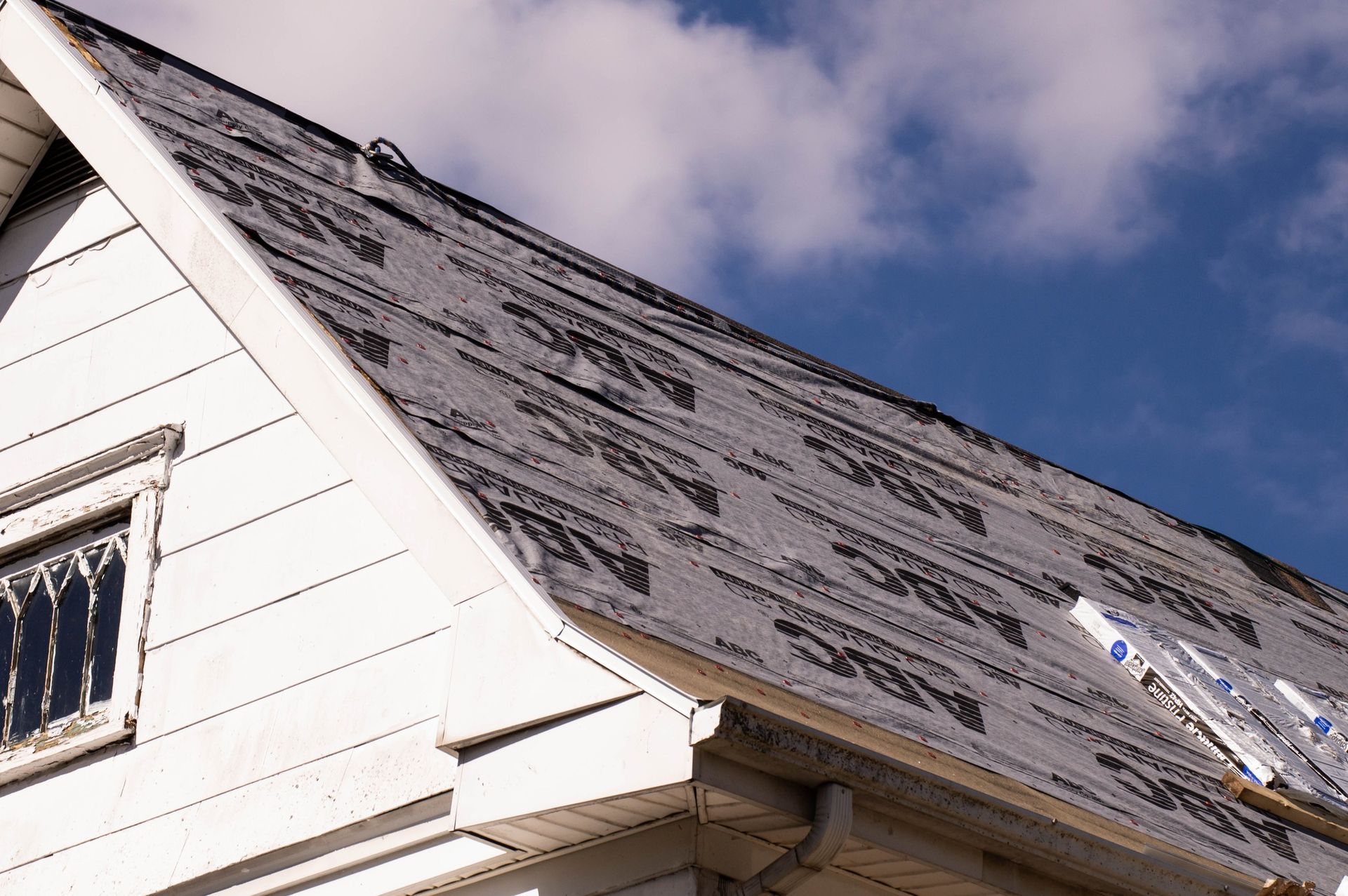 A white house roof partially covered with black roofing underlayment, blue sky and clouds in the background.