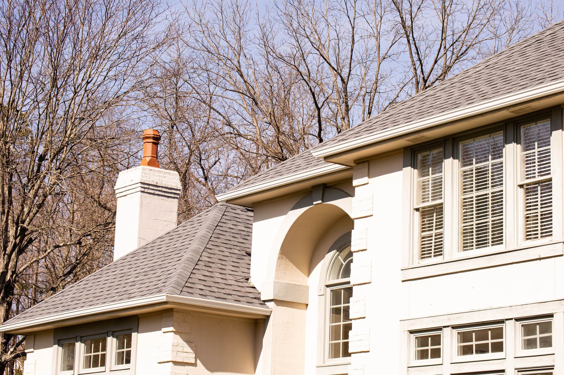Building exterior with gray roof, chimney, and arched window. Pale wall, bare trees.