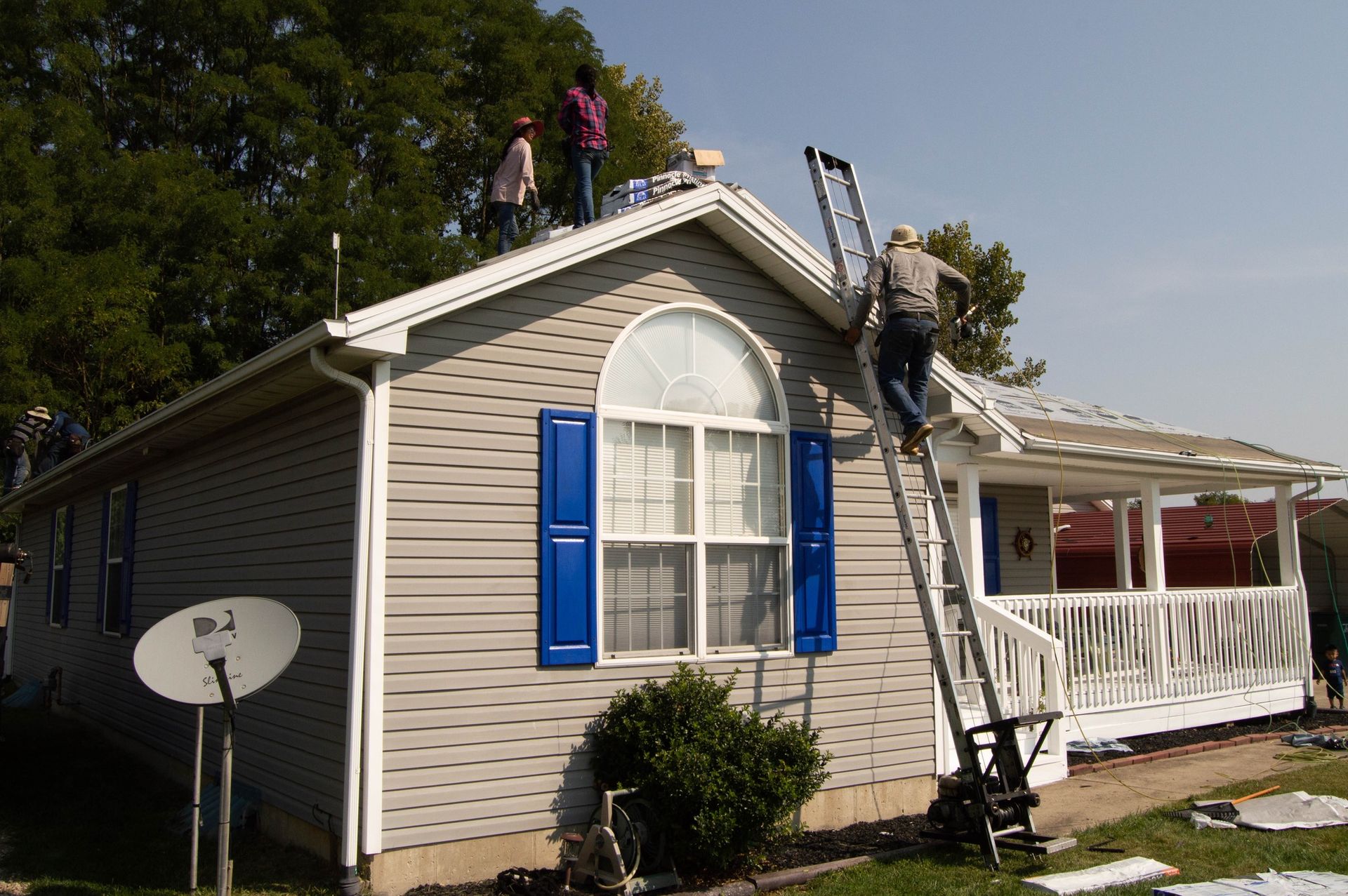 Three people on a roof with a person climbing a ladder. House has blue shutters and a satellite dish.