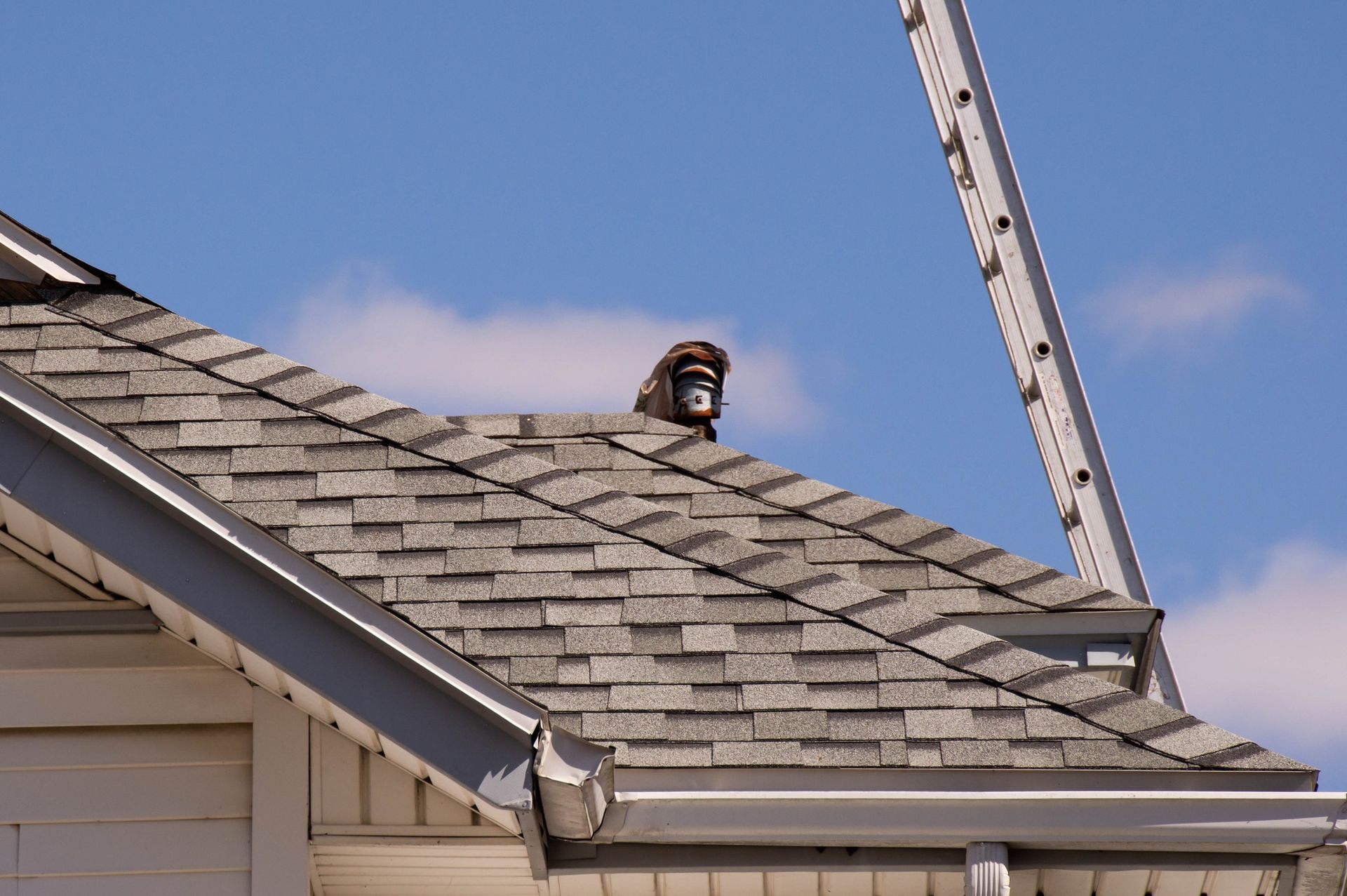 Roof of a house with gray shingles, gutters, and a security camera. A ladder leans against the roof.