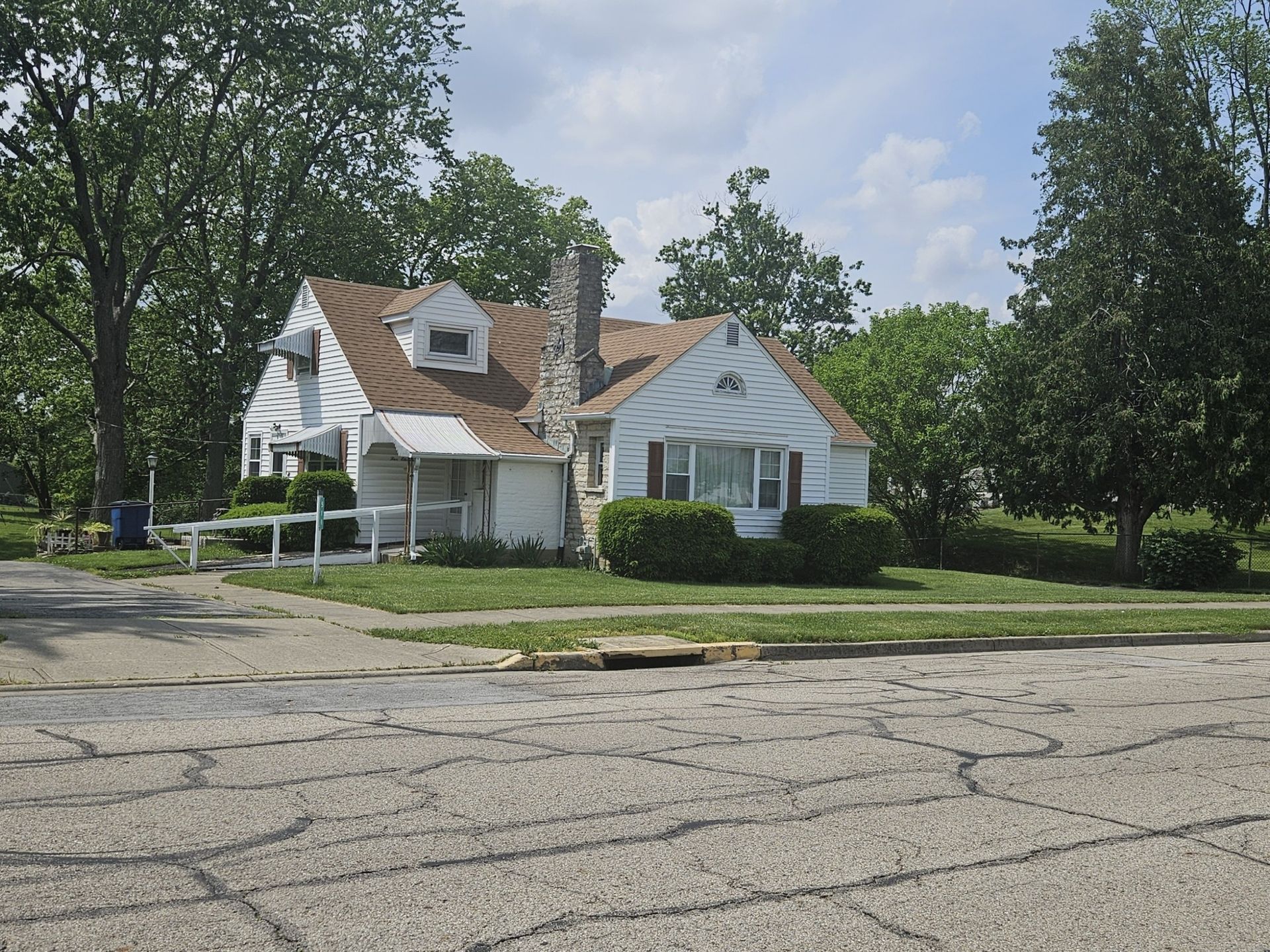 White house with brown roof, shrubs, and chimney on a cracked street, surrounded by green trees.