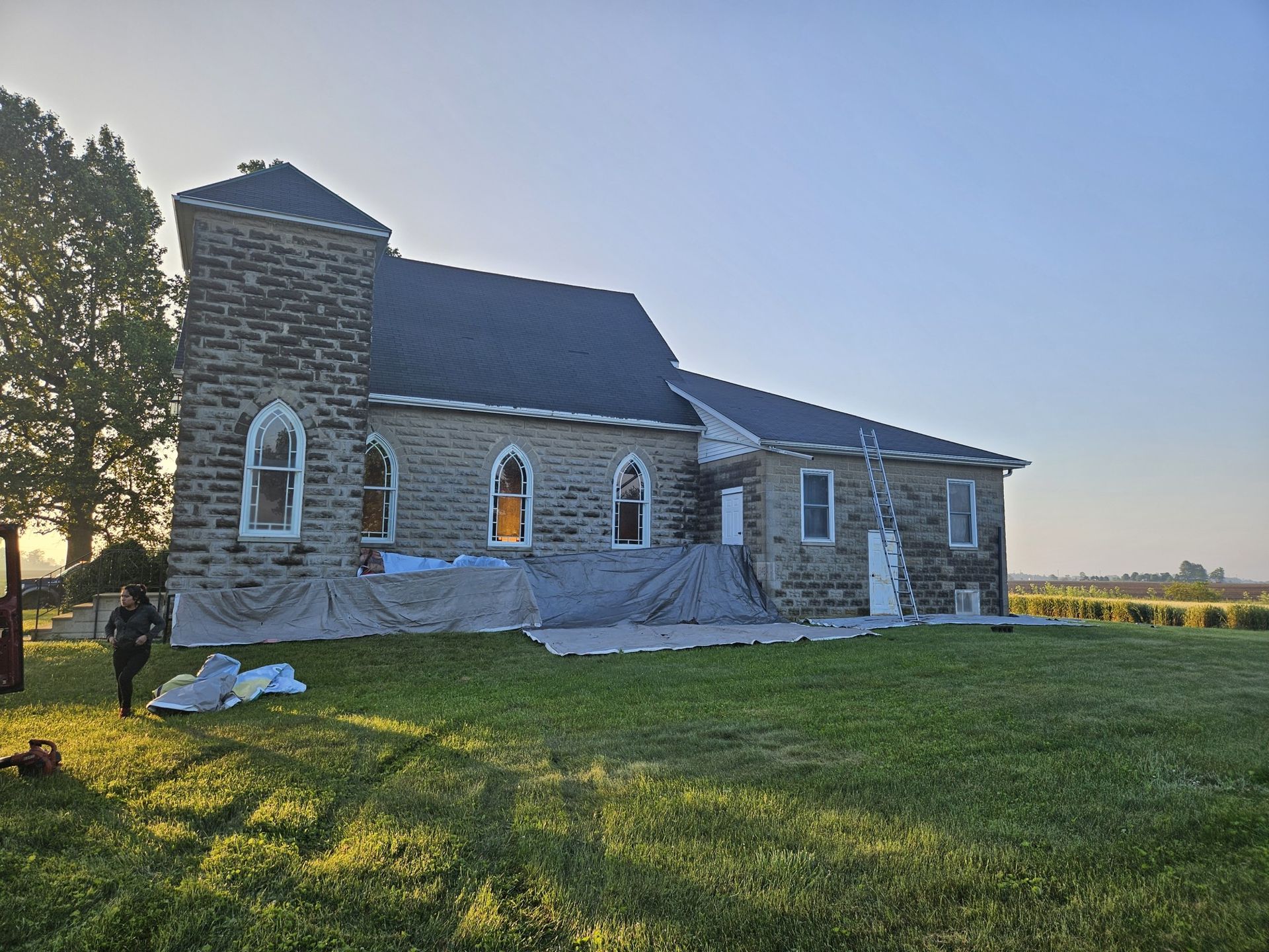 Stone church with black roof and white arched windows, set in a grassy field. A person stands nearby.