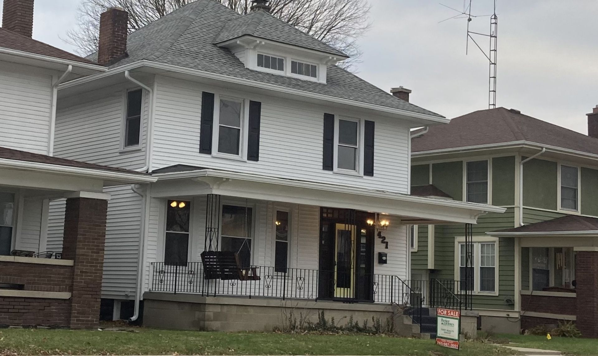 White two-story house with black shutters, porch swing, and green house next door under a cloudy sky.