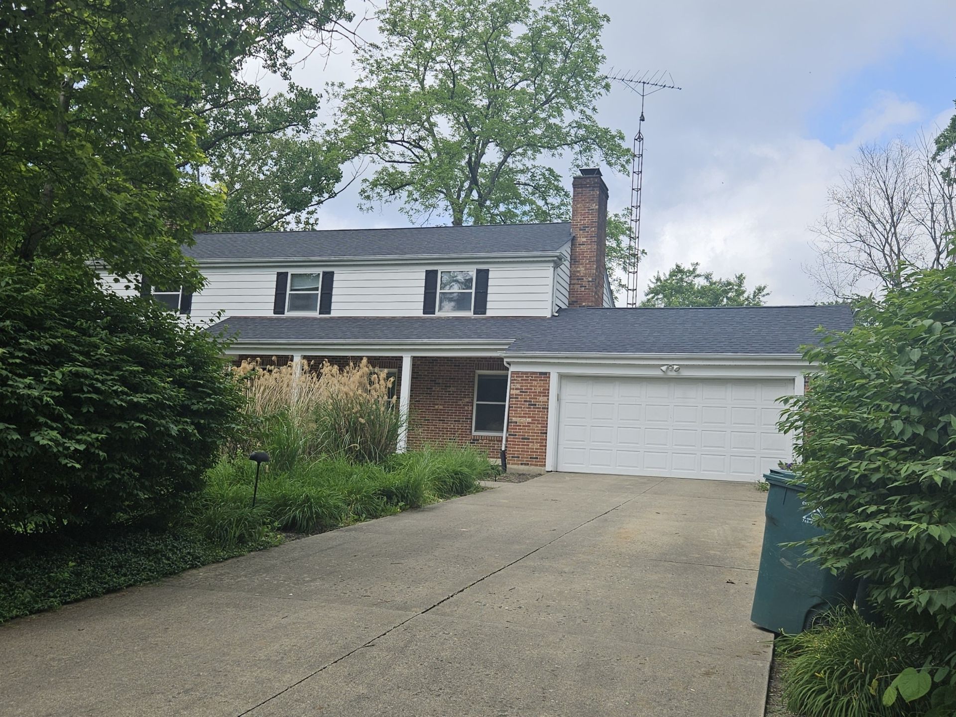 Two-story white house with brick accents, attached garage, and a long driveway. Overgrown landscaping.