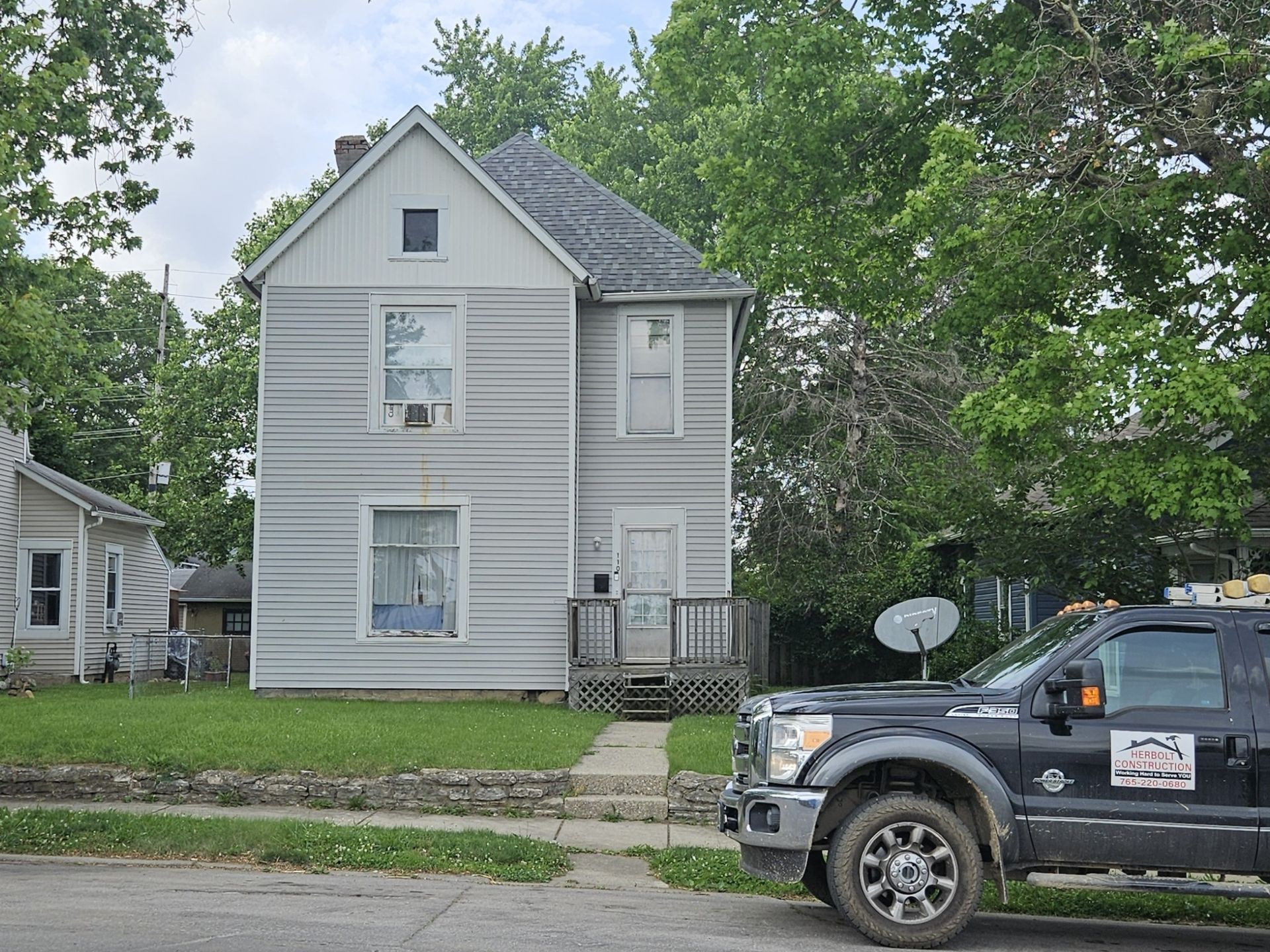 Two-story gray house with small porch, next to a black truck, green lawn, and trees.