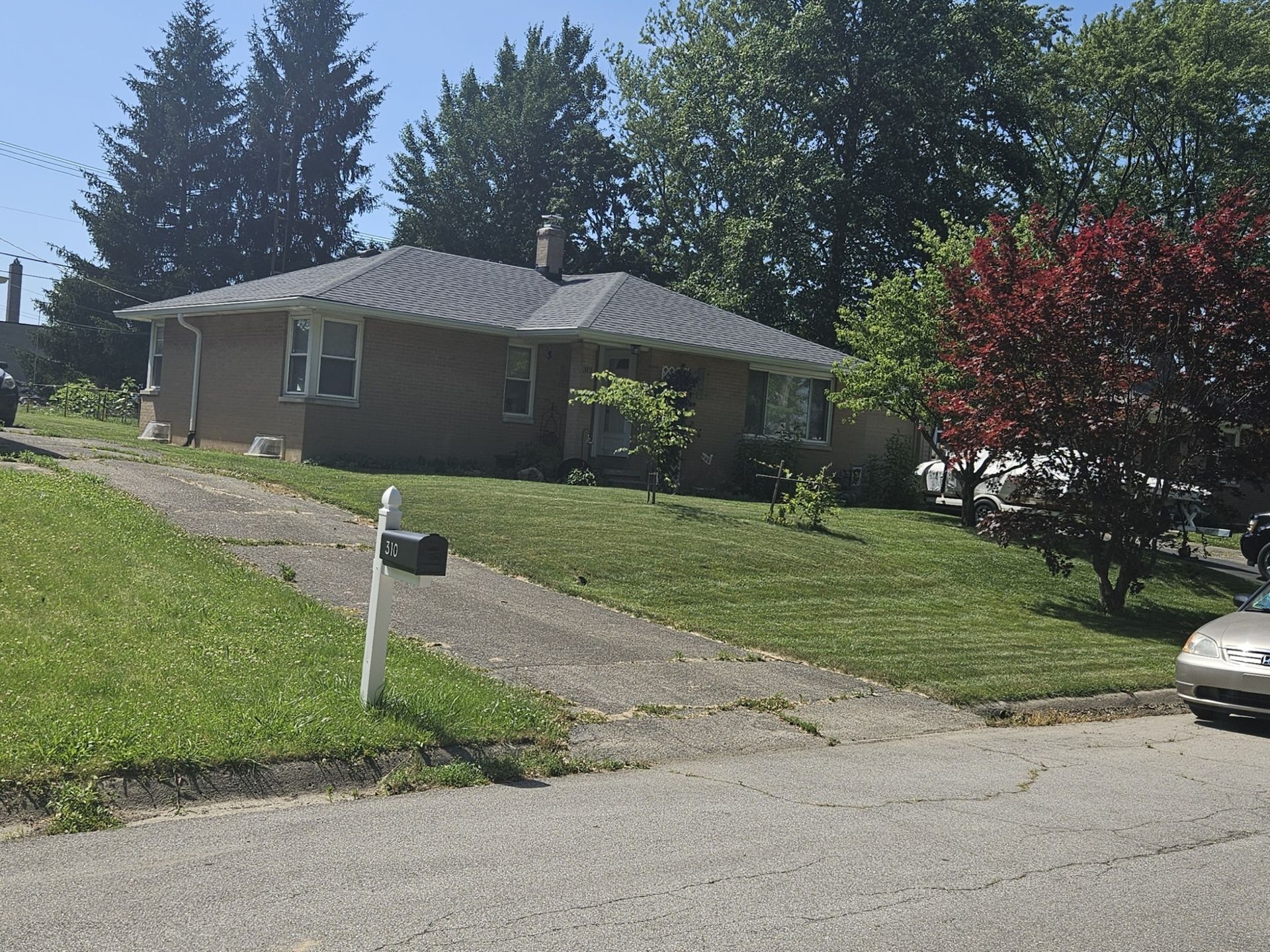 A small brick house with a driveway and mailbox sits on a grassy hill; trees surround the house.