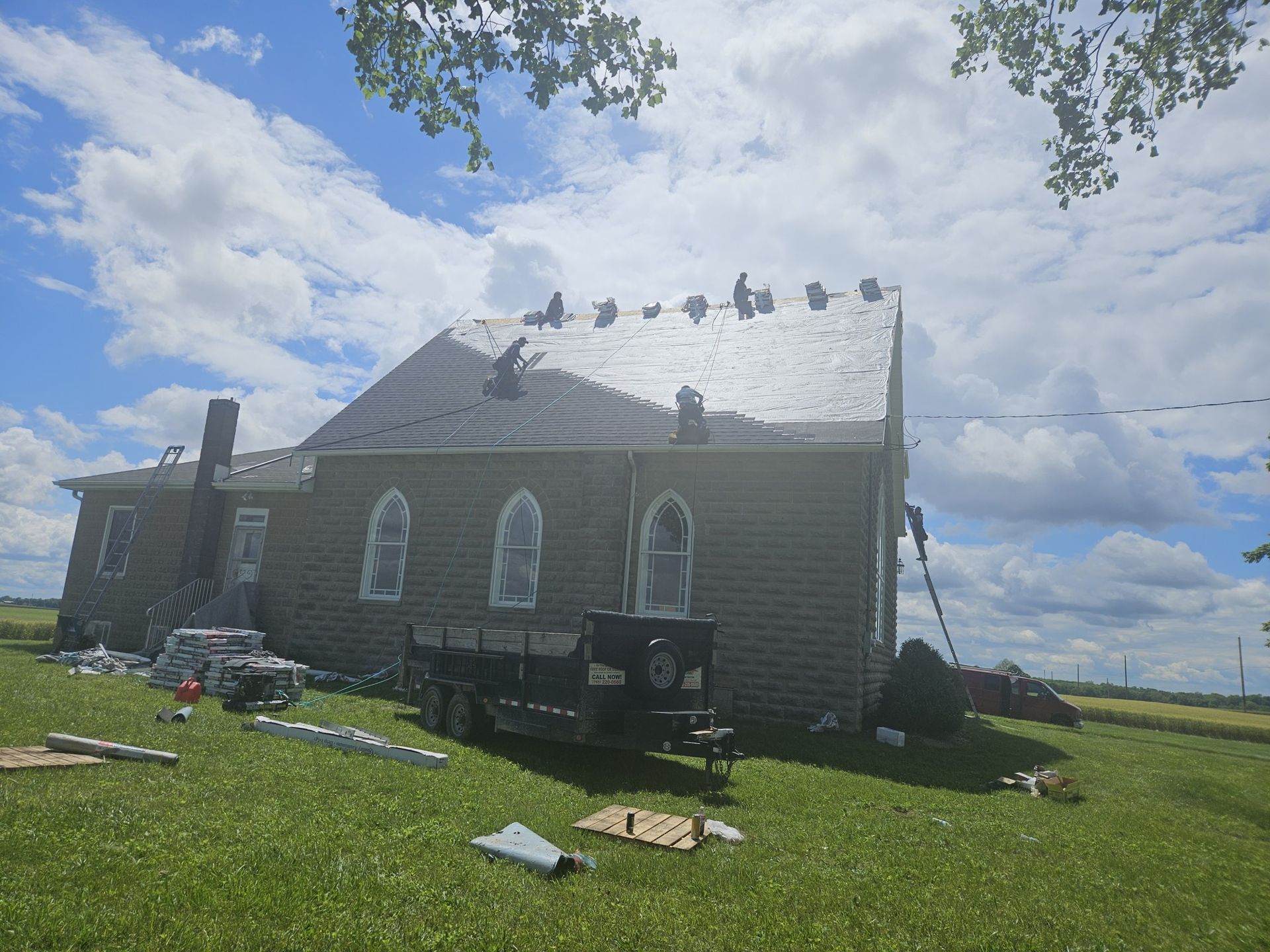 Church with damaged roof, debris on the ground, and a trailer parked outside on a green grassy field under a cloudy sky.