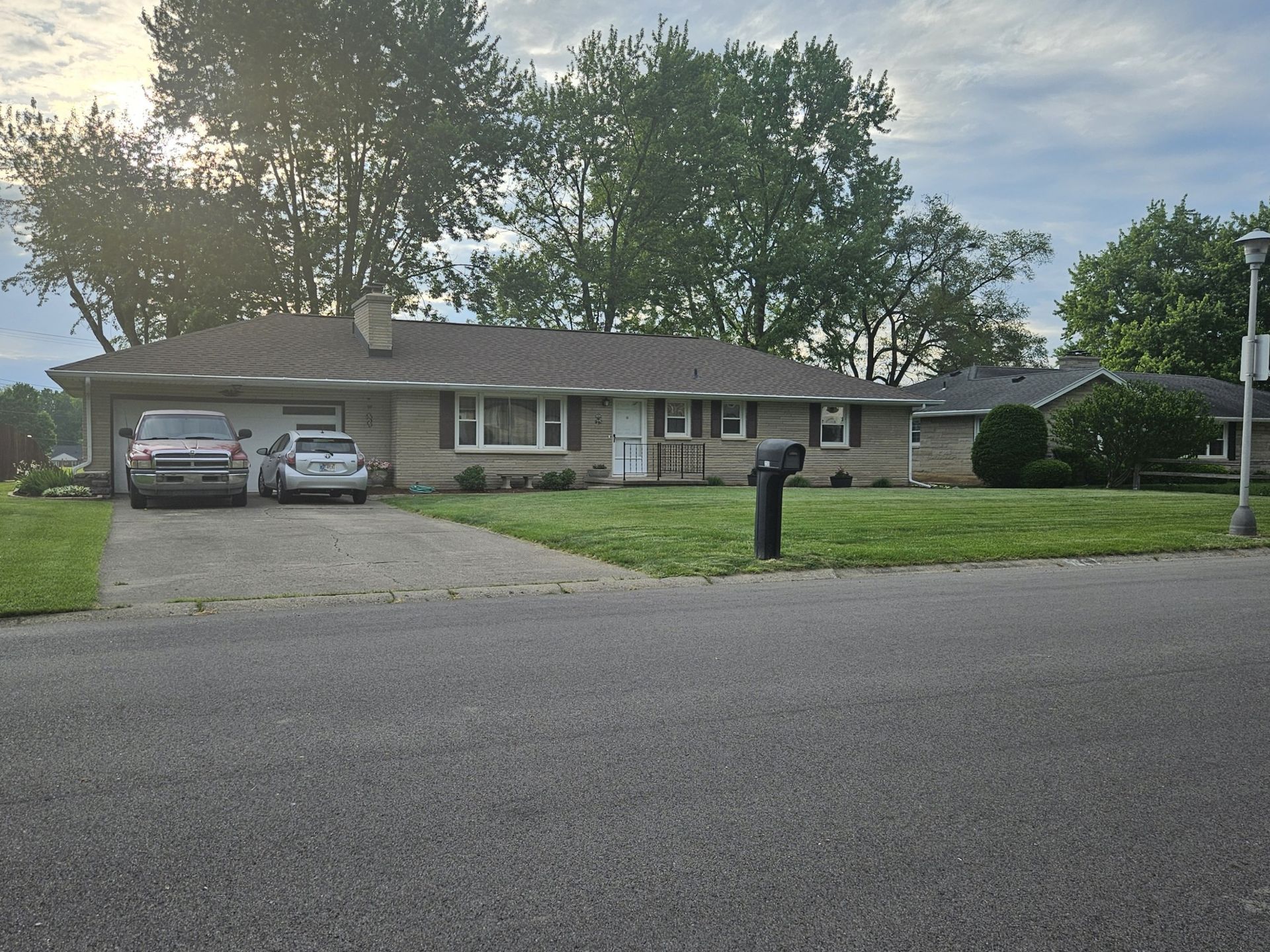 A ranch-style house with a driveway. Two cars are parked in the driveway. Trees line the background.