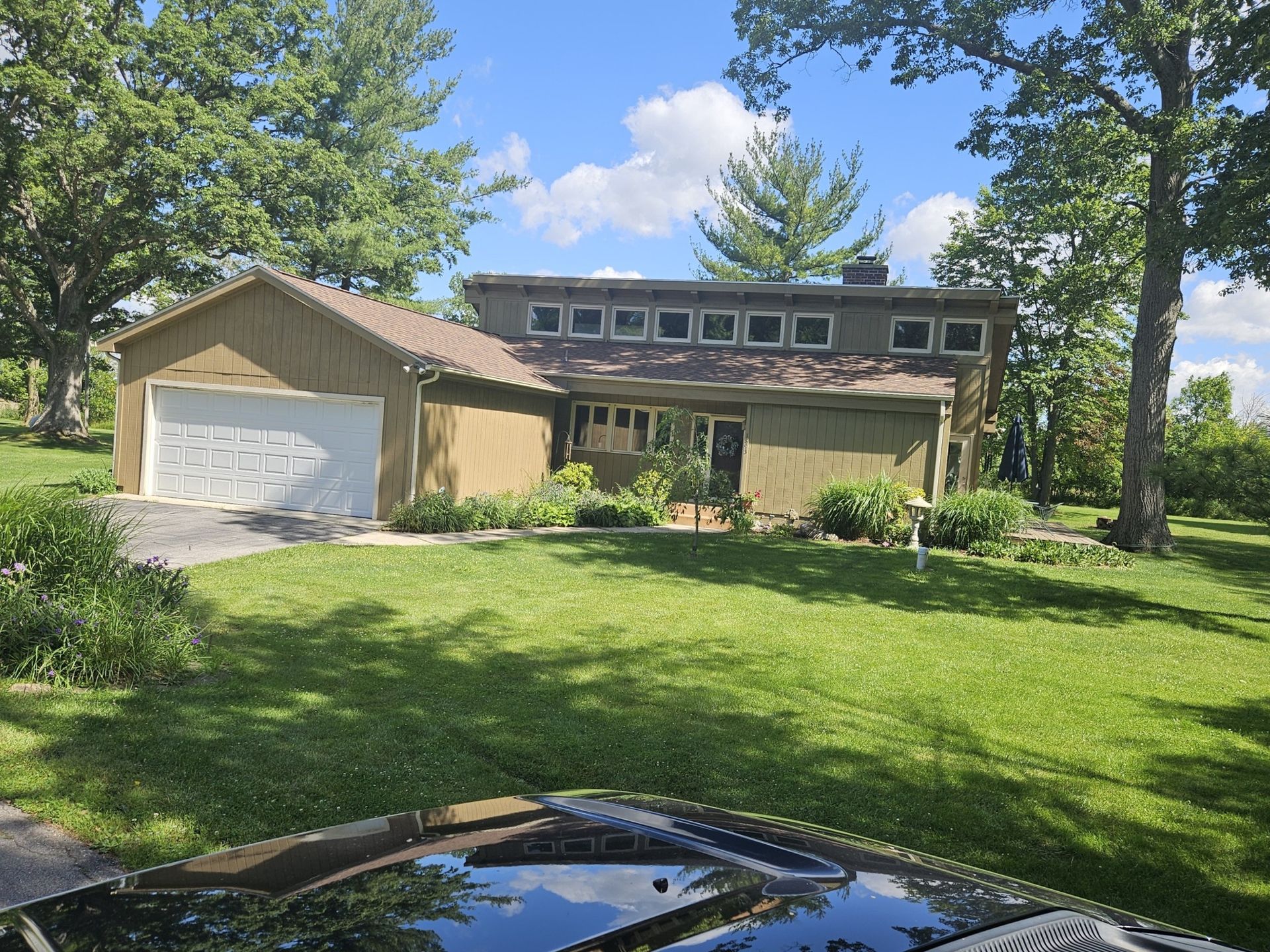 House with tan siding, attached garage, green lawn, trees, and blue sky.