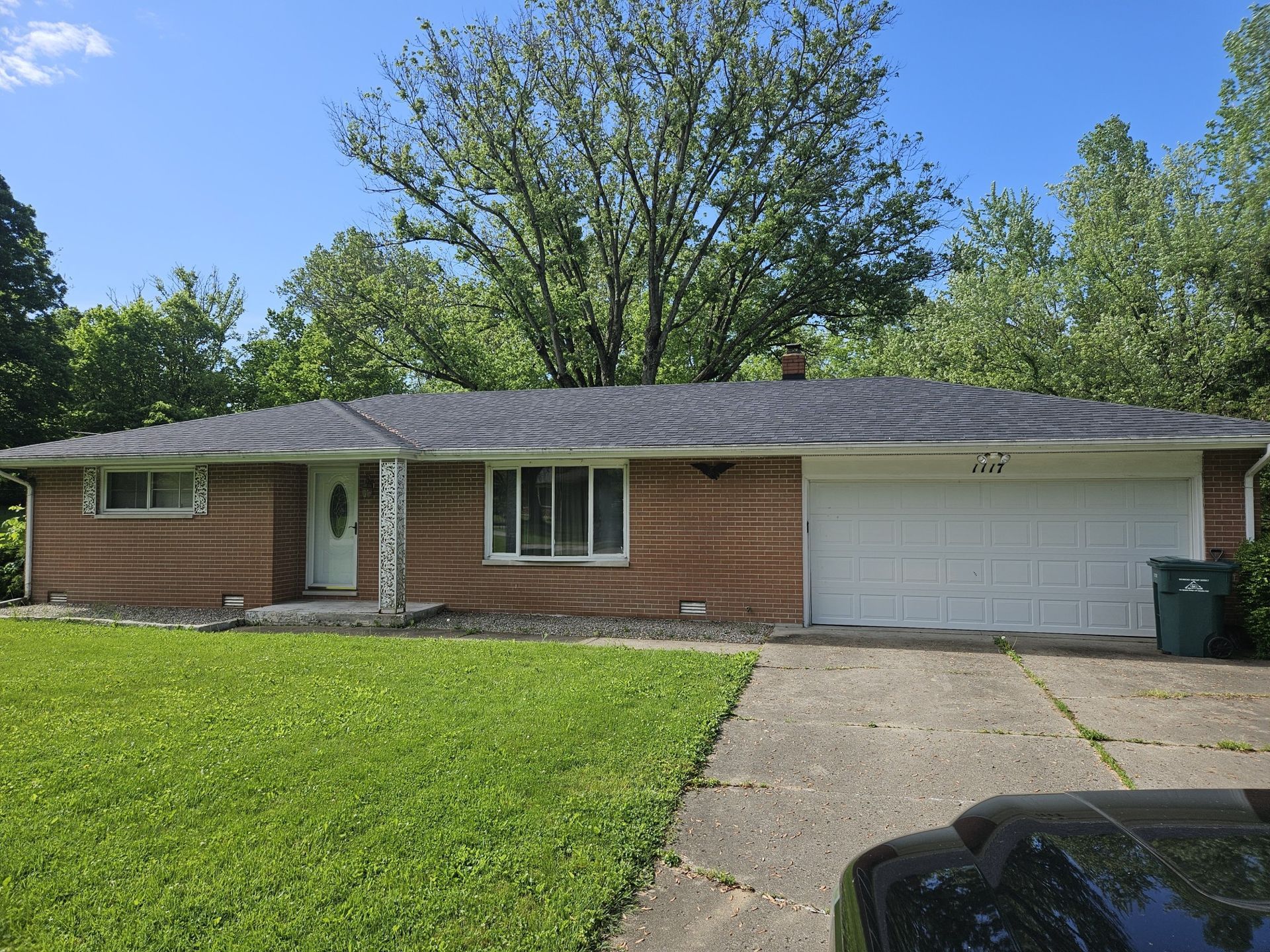 Brick ranch-style house with a gray roof and white garage door; green lawn and trees in the background.
