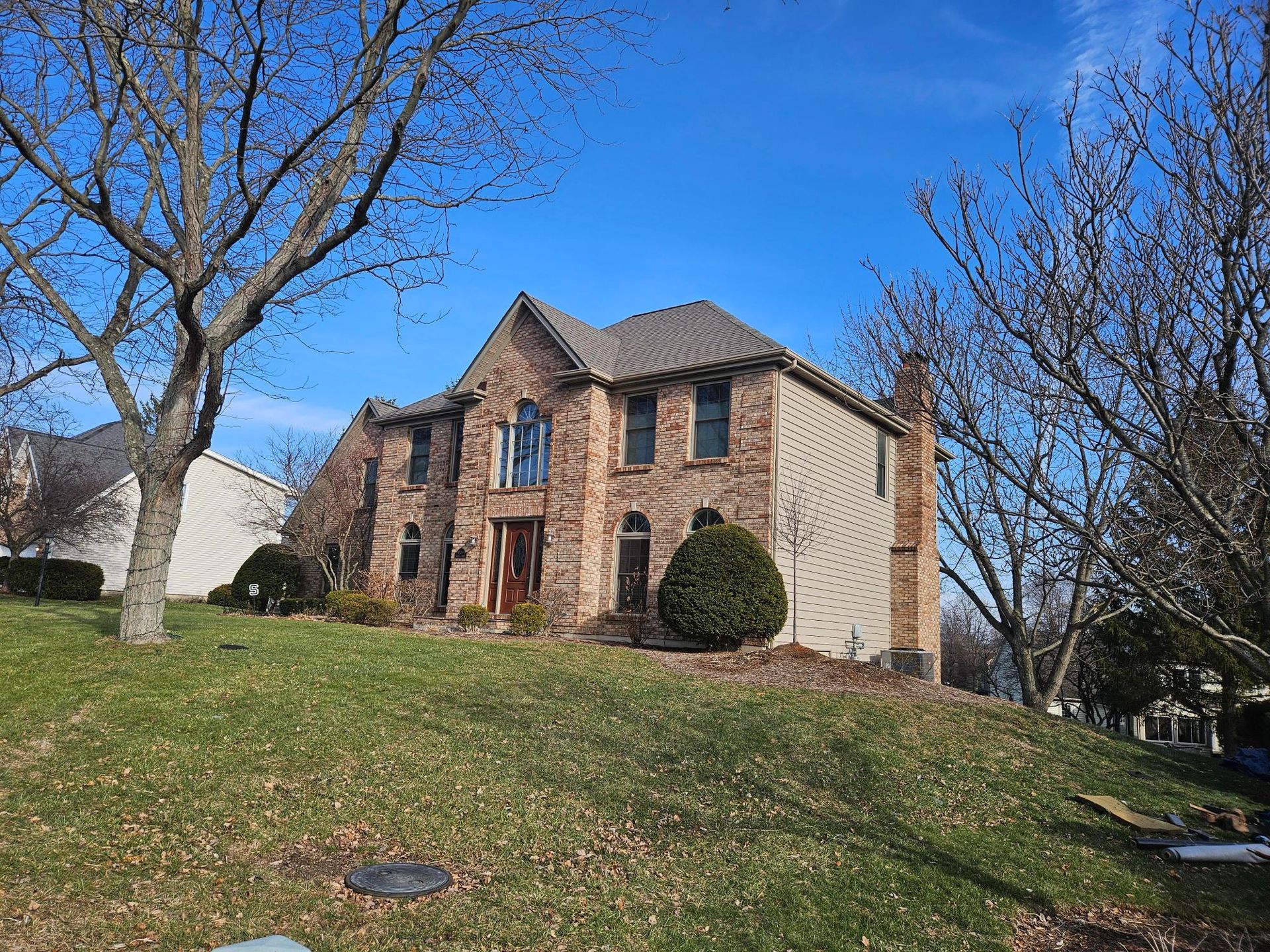 Two-story brick house with a brown roof and a green lawn under a blue sky. Bare trees flank the house.