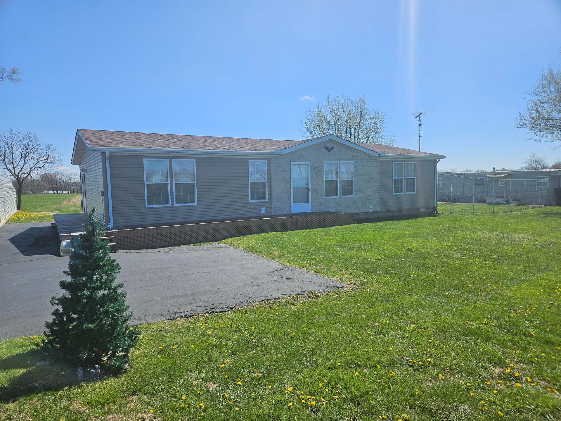 Ranch-style house with gray siding and brown roof on a grassy lot with a paved driveway, under a blue sky.