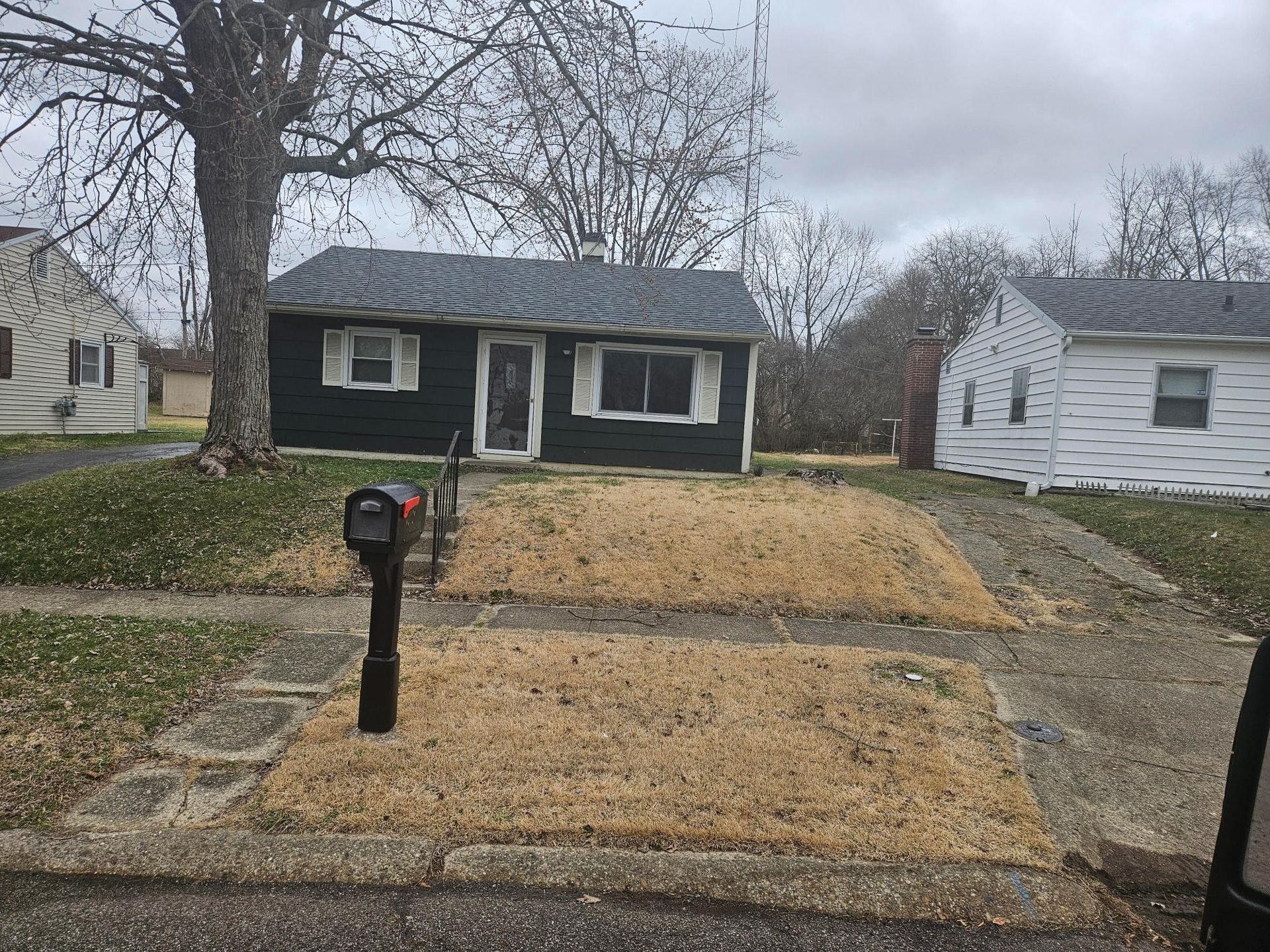 Small dark house with brown, dead grass in front. Black mailbox in foreground. Overcast day.
