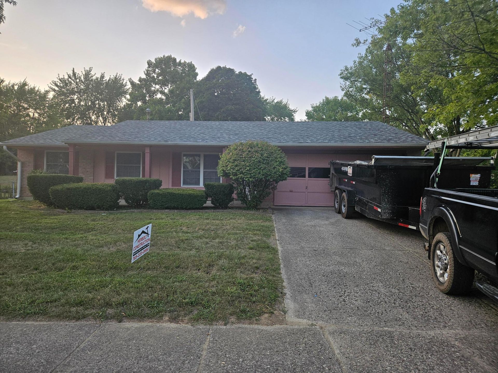 Pink ranch house with gravel driveway. Black trailer and truck parked. Green lawn and bushes in front.
